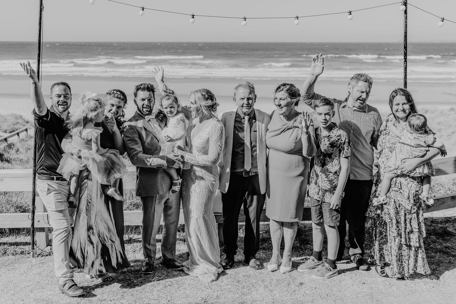 A wedding party gathers for a group photo on a beach with string lights overhead and ocean waves in the background. Papamoa photographer