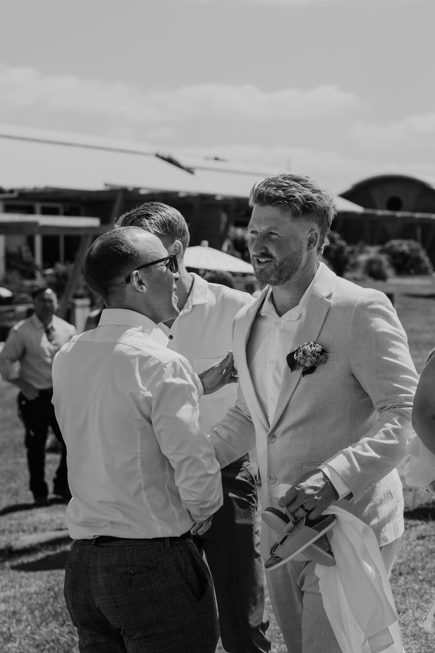 Groom in white suit with boutonniere laughing with guests outdoors at sunny wedding celebration.