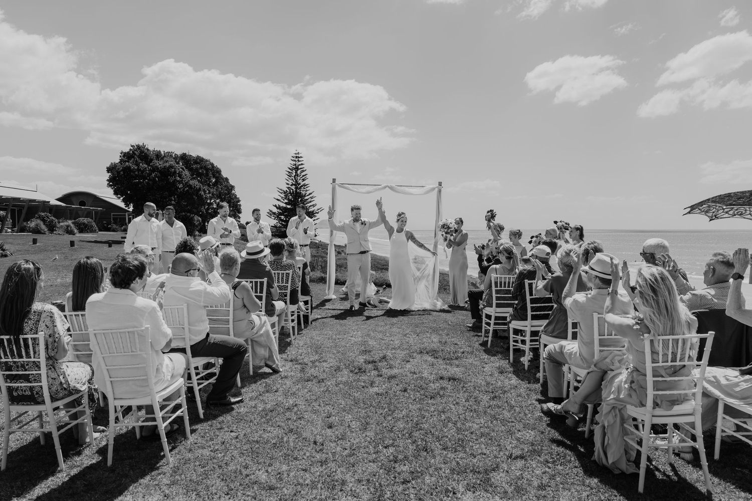 Black and white outdoor wedding ceremony by the ocean with guests seated in white chairs on a sunny day.