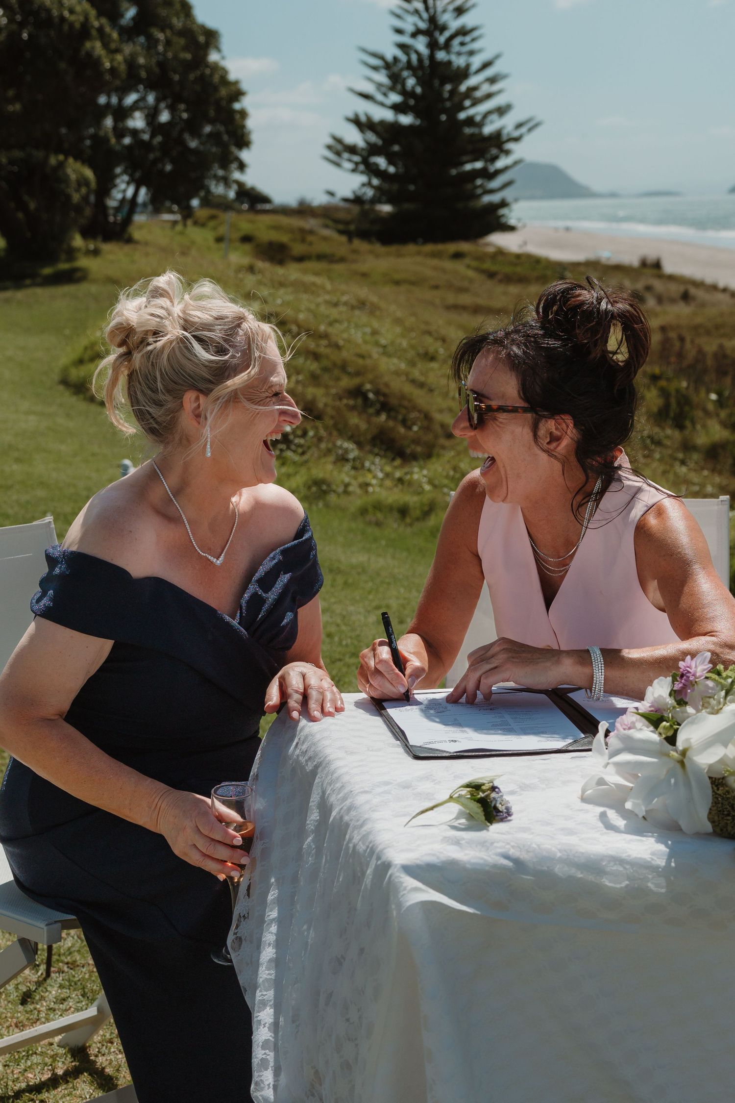 Two women laugh together at an outdoor coastal wedding table, one in navy off-shoulder dress, other in pink signing documents.