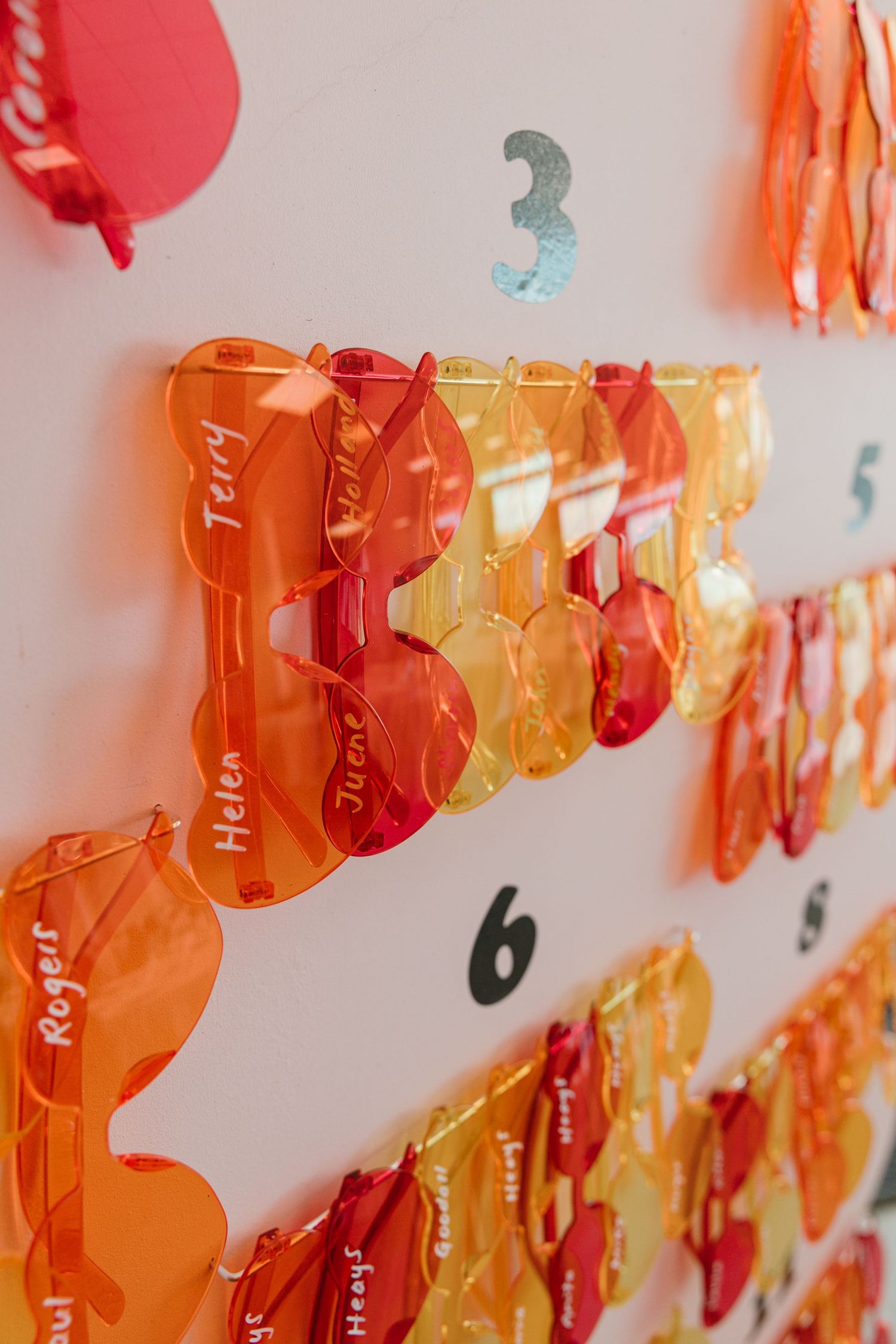 Colorful transparent acrylic sunglasses with names written on them displayed on a white wall with numbers.