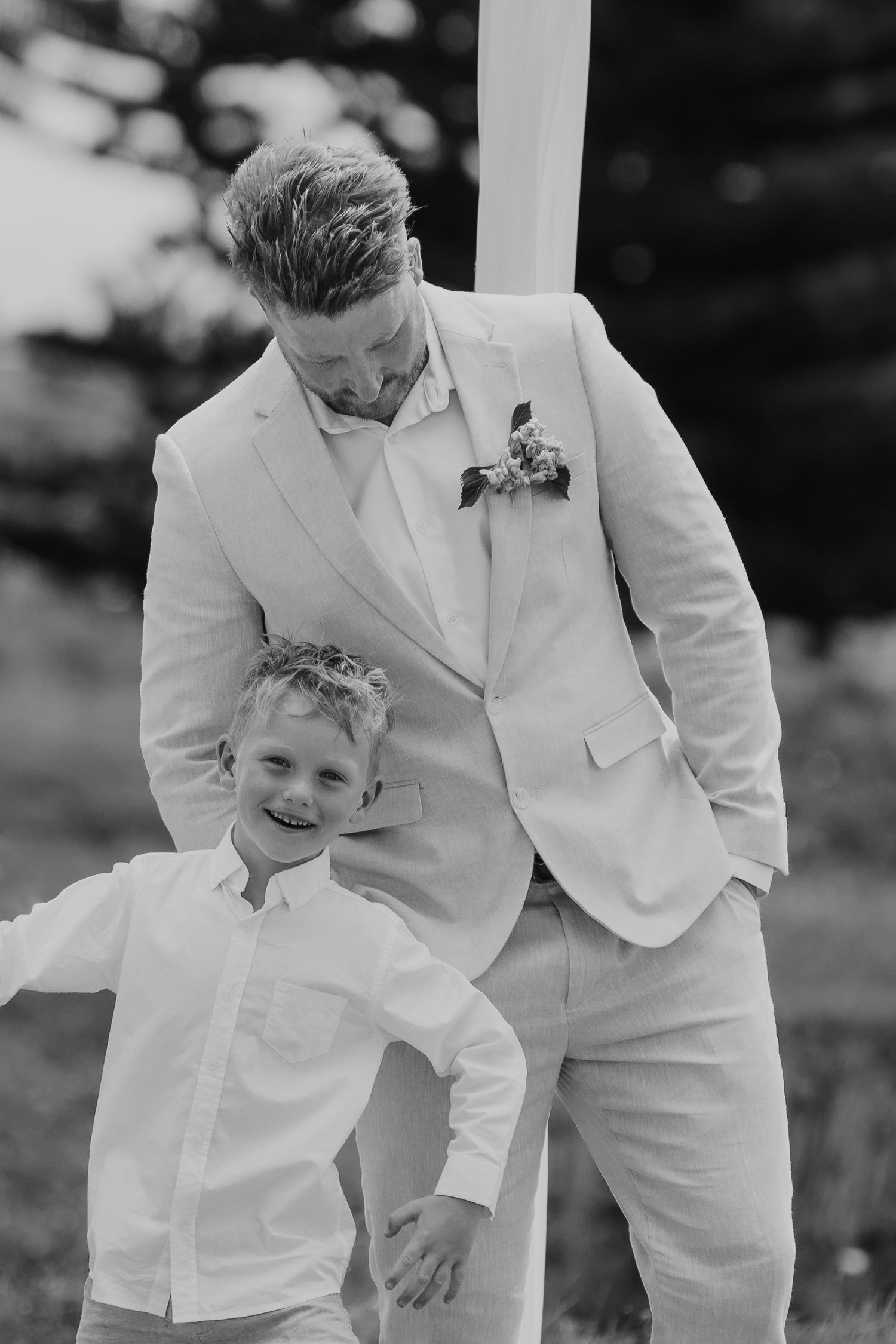 Black and white photo of a groom in a white suit smiling down at a young boy in a white shirt at a wedding.
