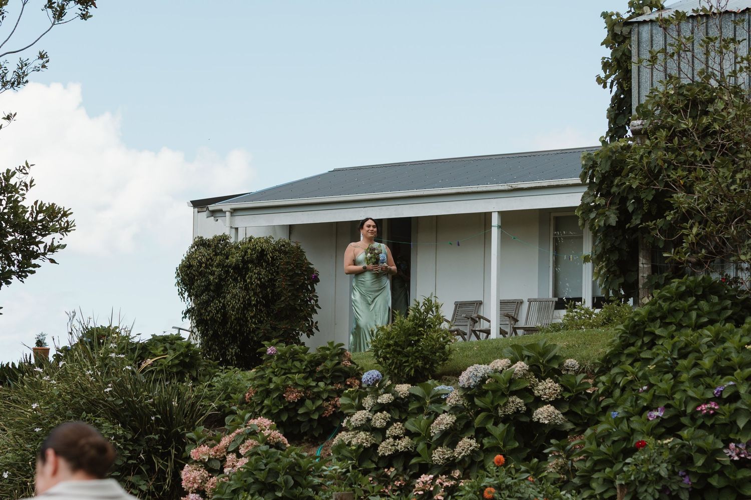 Bridesmaid in sage green dress holding bouquet stands on elevated garden terrace beside white country homestead.
