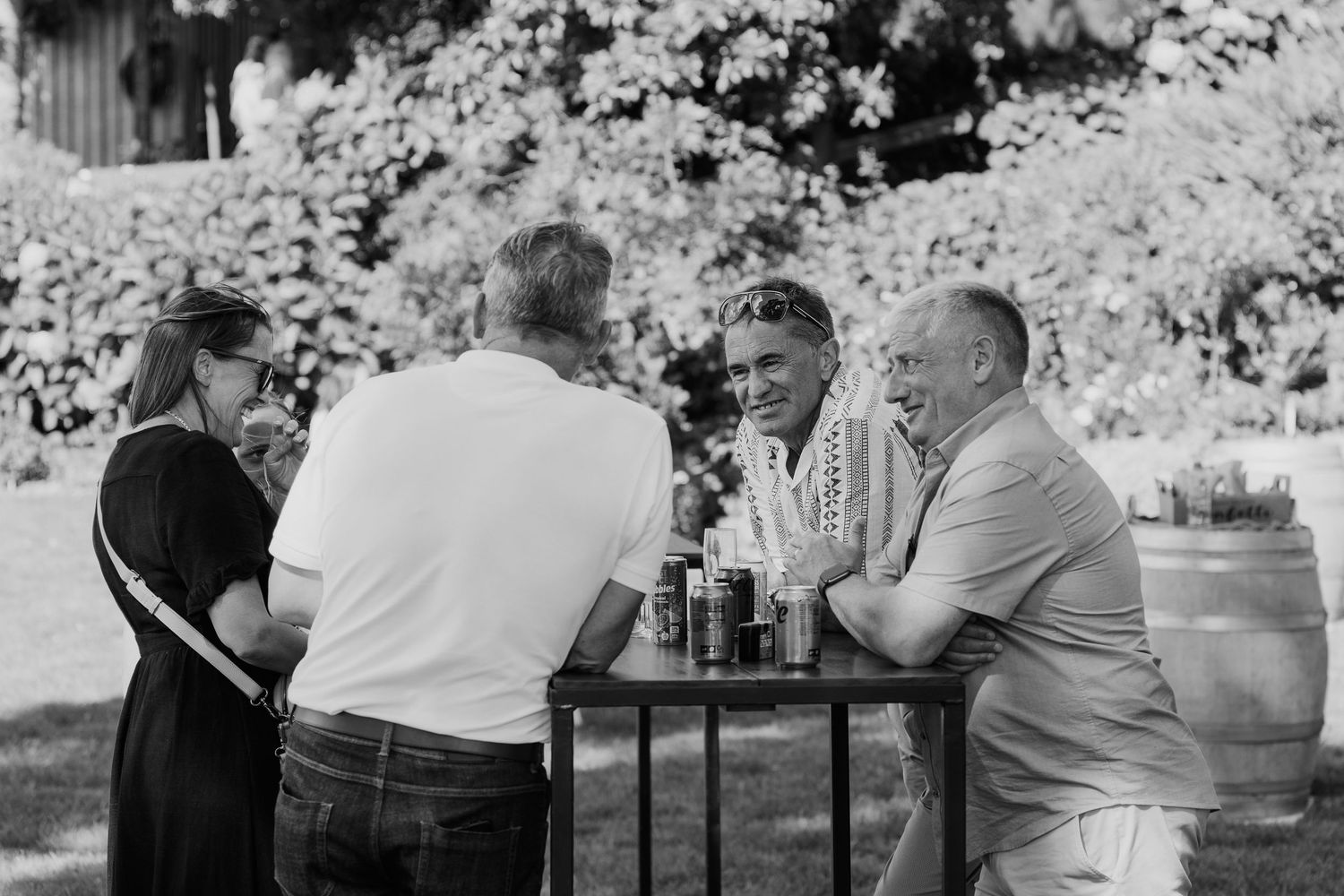 Black and white photo of four adults socializing around an outdoor table with drinks in a garden setting.