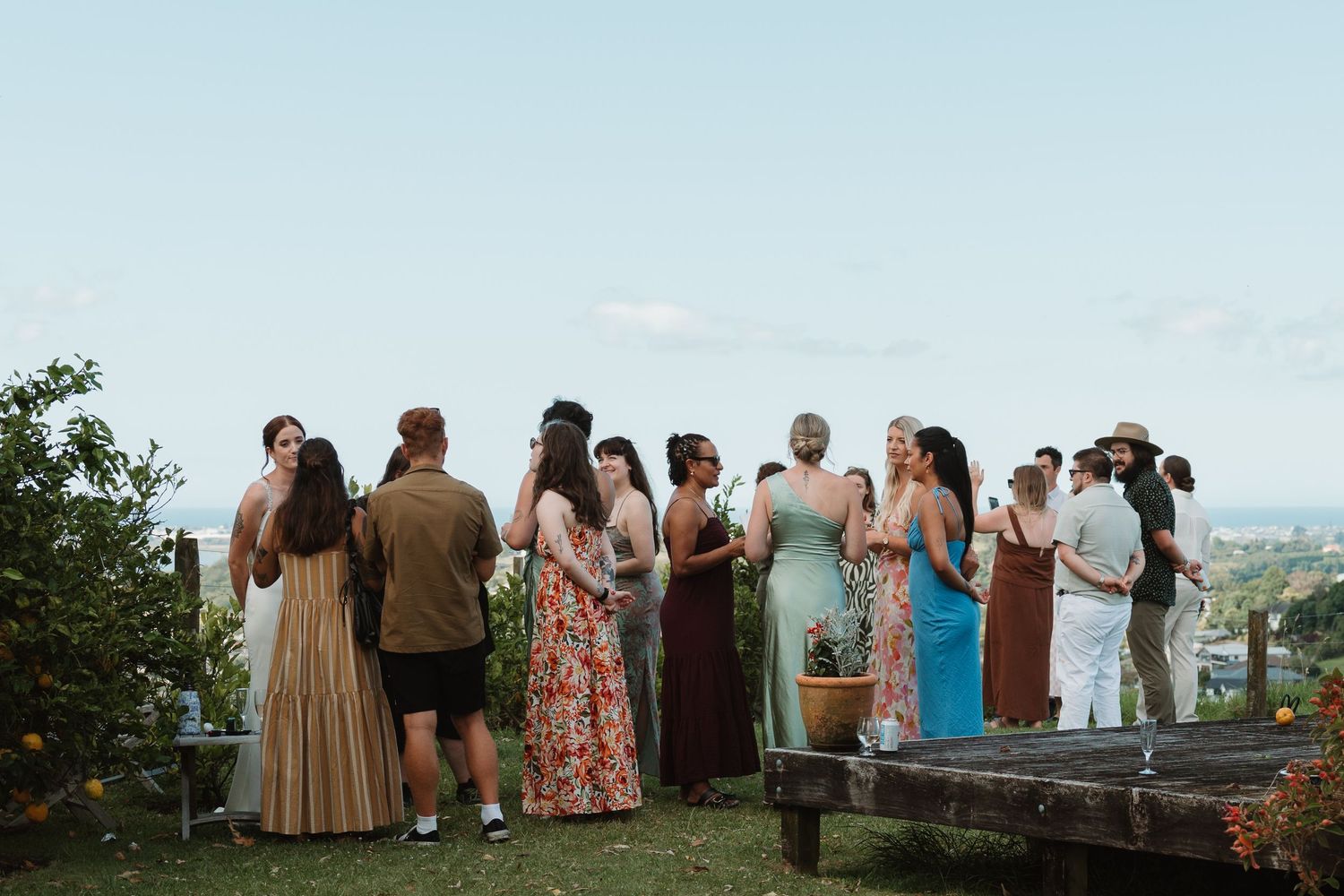 Guests mingling outdoors at a hilltop garden party with scenic views, dressed in colorful summer attire.