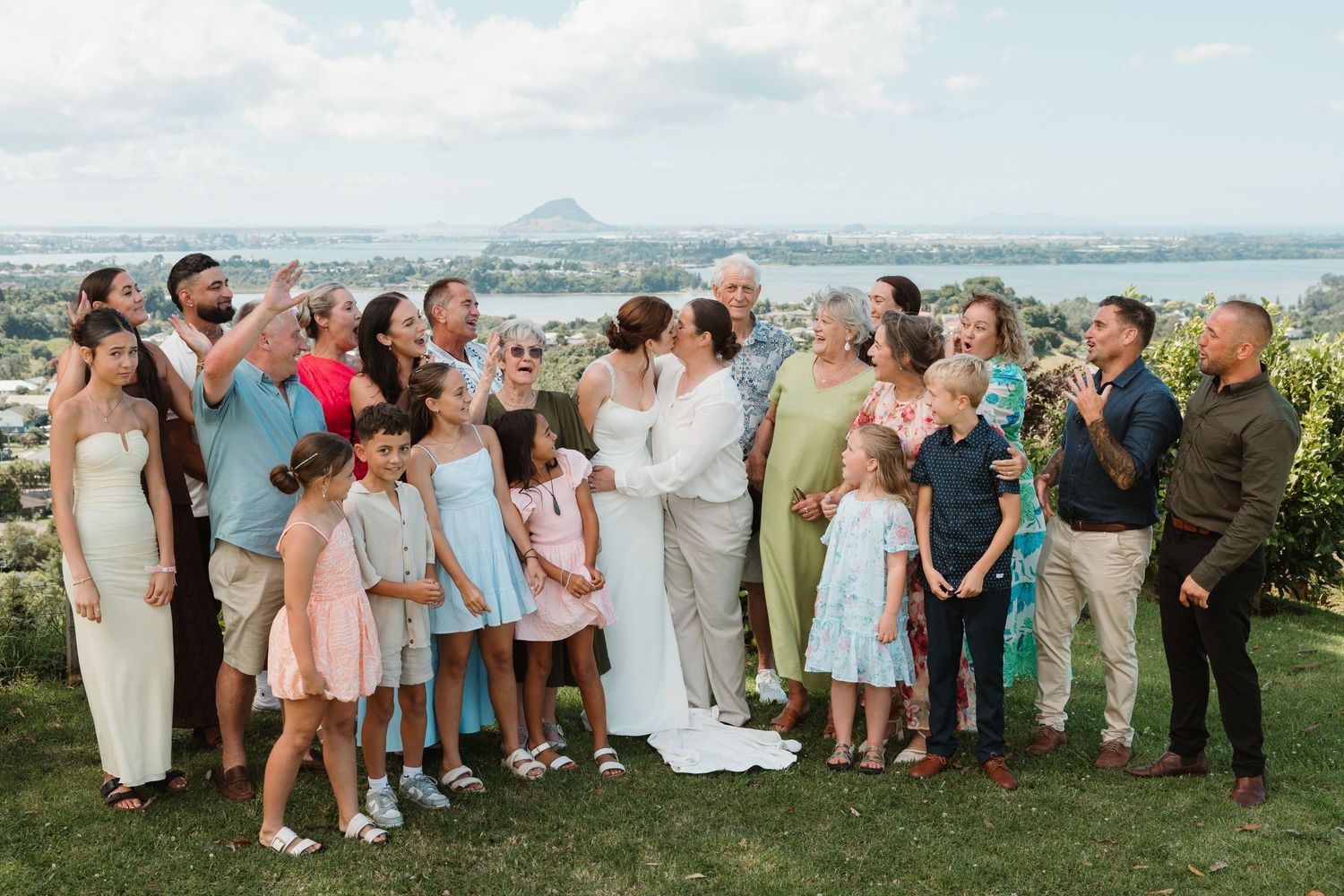 Wedding group photo outdoors with couple kissing surrounded by family and children, scenic water and mountain views.