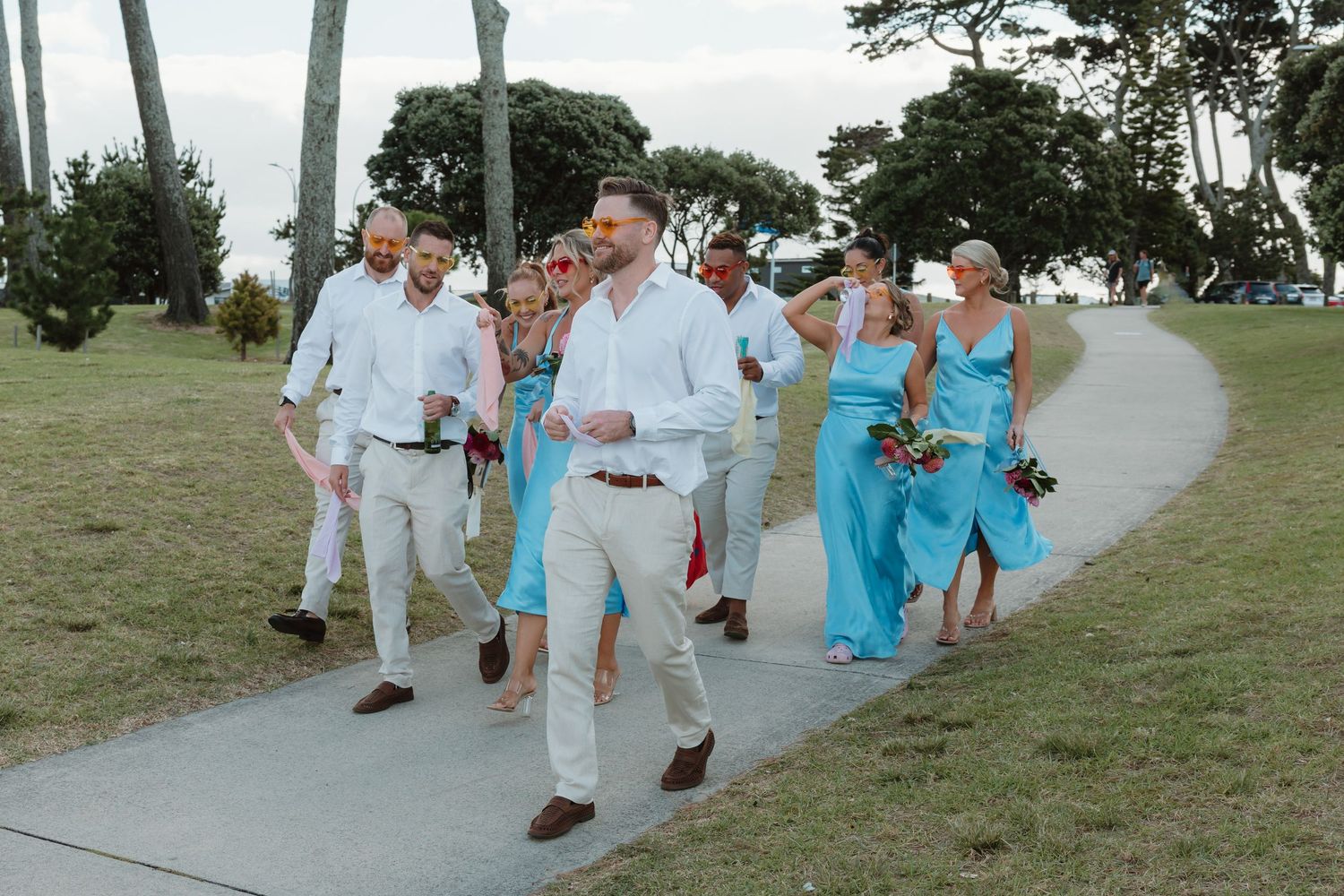 Wedding party walks on path, bridesmaids in turquoise dresses, groomsmen in white shirts and tan pants, outdoor ceremony.