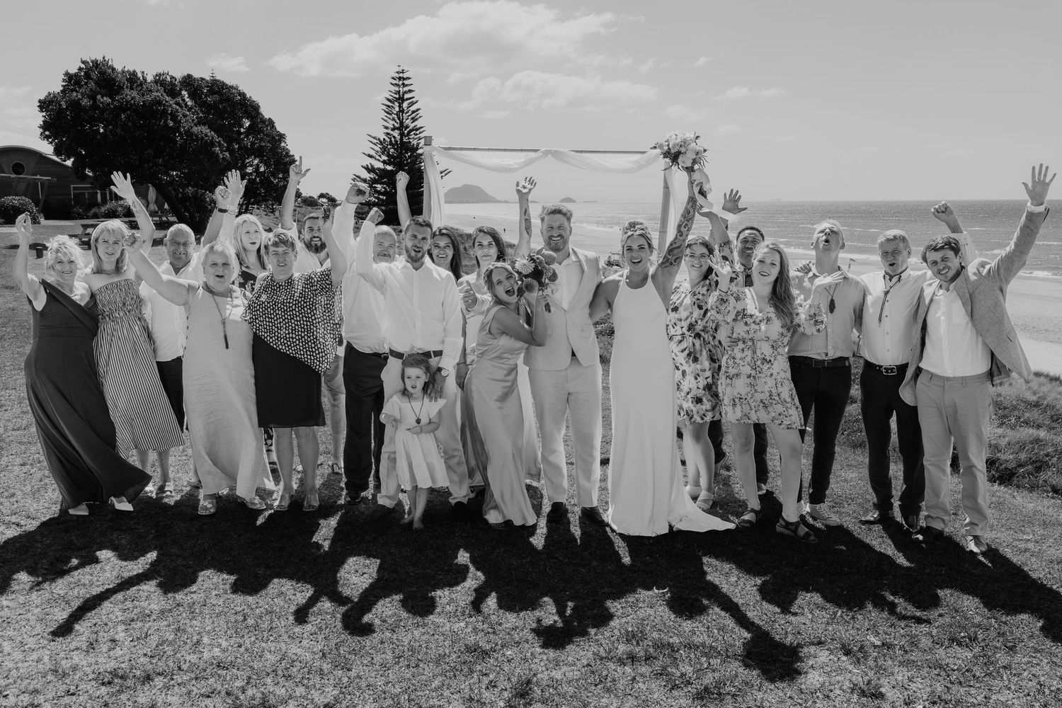 Black and white wedding photo of a joyful group celebrating outdoors near the ocean, with bride and groom surrounded by guests.