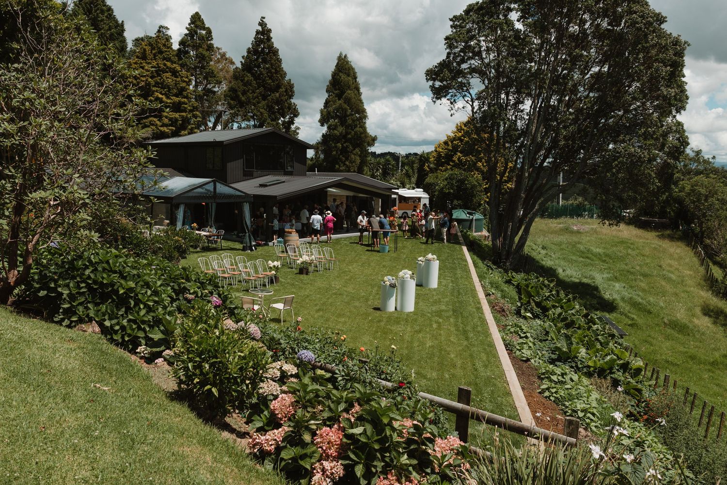 Scenic outdoor wedding venue with lush green gardens, hydrangeas, and guests gathered near a dark modern house.