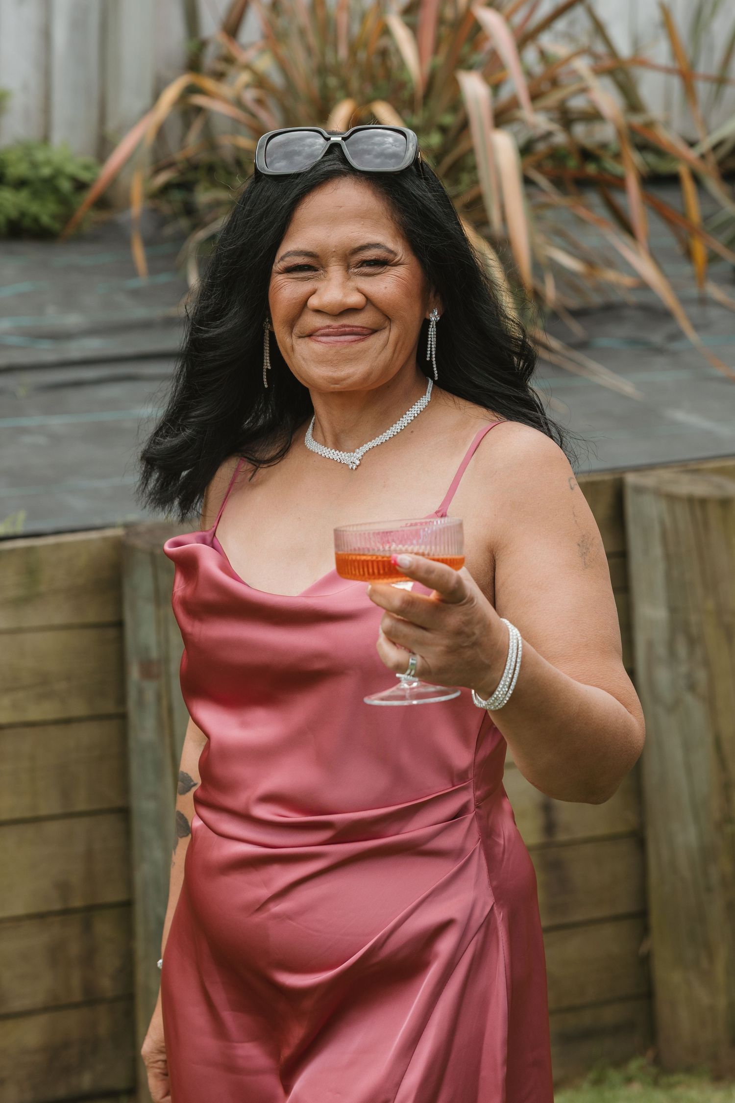 A person in a pink satin dress holds up a cocktail glass while posing against a wooden fence outdoors.