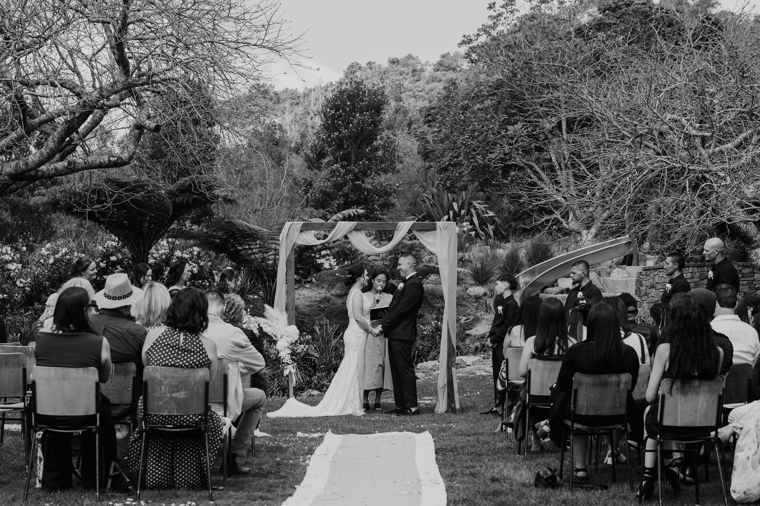 A panoramic black and white photograph shows guests seated outdoors at a wedding ceremony under trees.