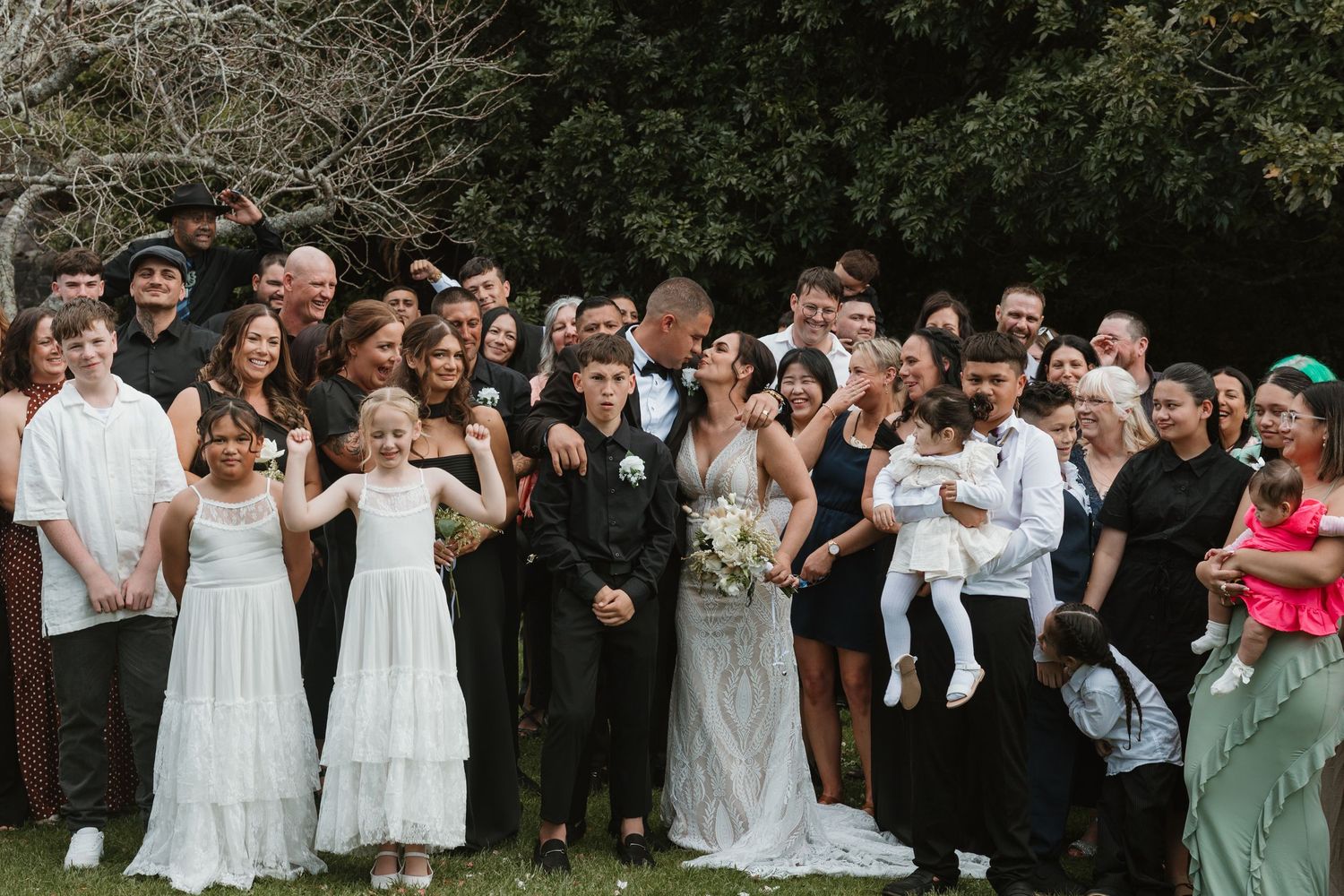 Wedding guests and wedding party stand in a row outdoors taking photos with their phones and cameras.