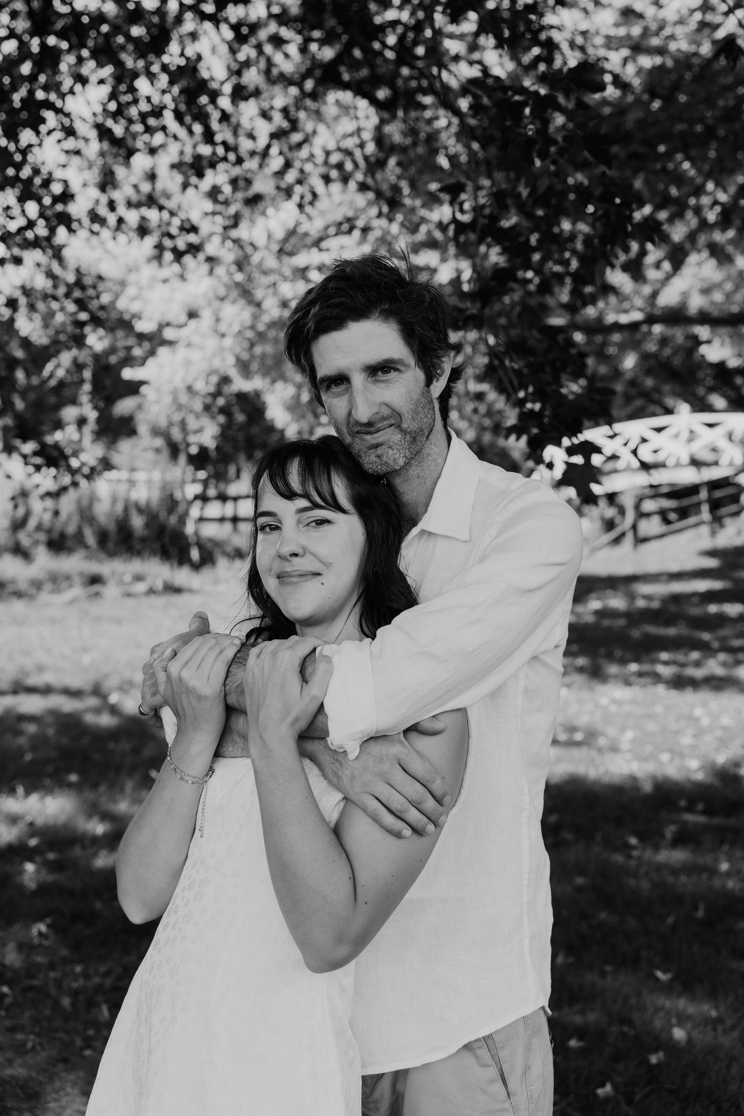 Black and white photo of a couple embracing outdoors, man in white shirt hugging woman in white dress among trees.