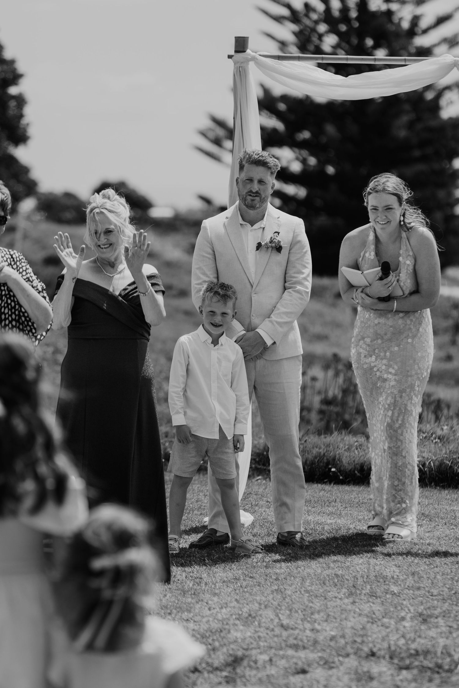 Black and white wedding ceremony photo of groom in linen suit with young boy and bridesmaid on a grassy outdoor venue.