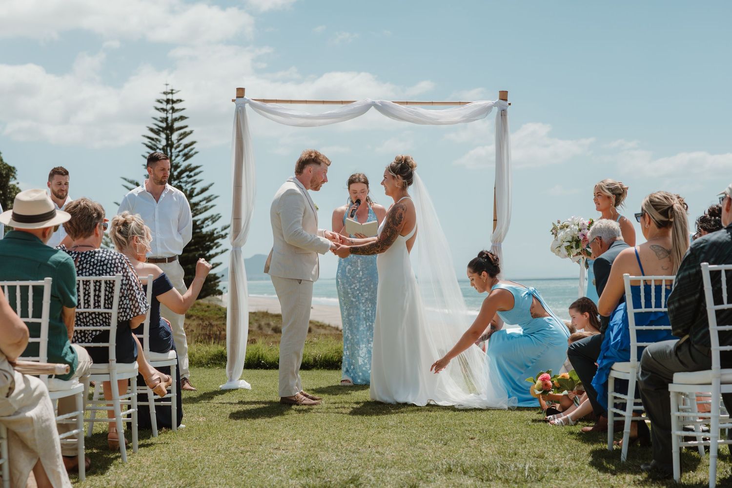 Outdoor beach wedding ceremony with couple exchanging vows under a white arch, guests seated on white chairs beside the ocean.