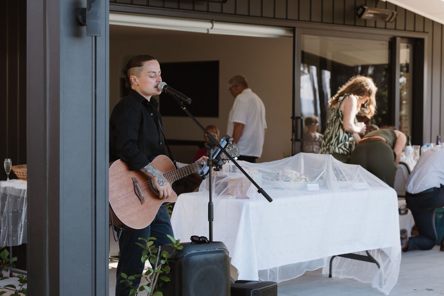 Musician playing acoustic guitar and singing at outdoor venue with guests dining in background.