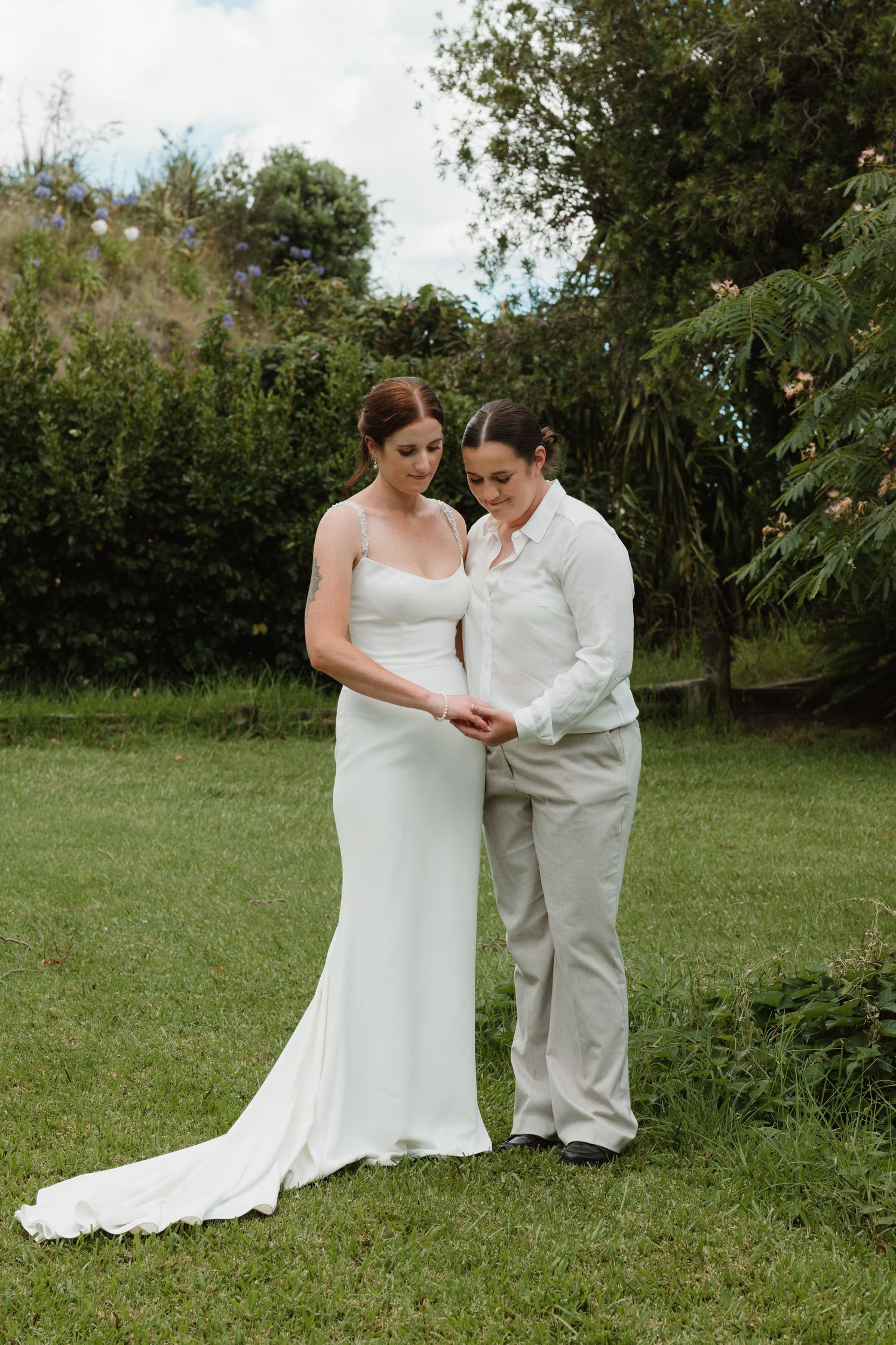 Two brides stand together on green lawn, one in white wedding gown, other in gray suit, sharing a tender moment.