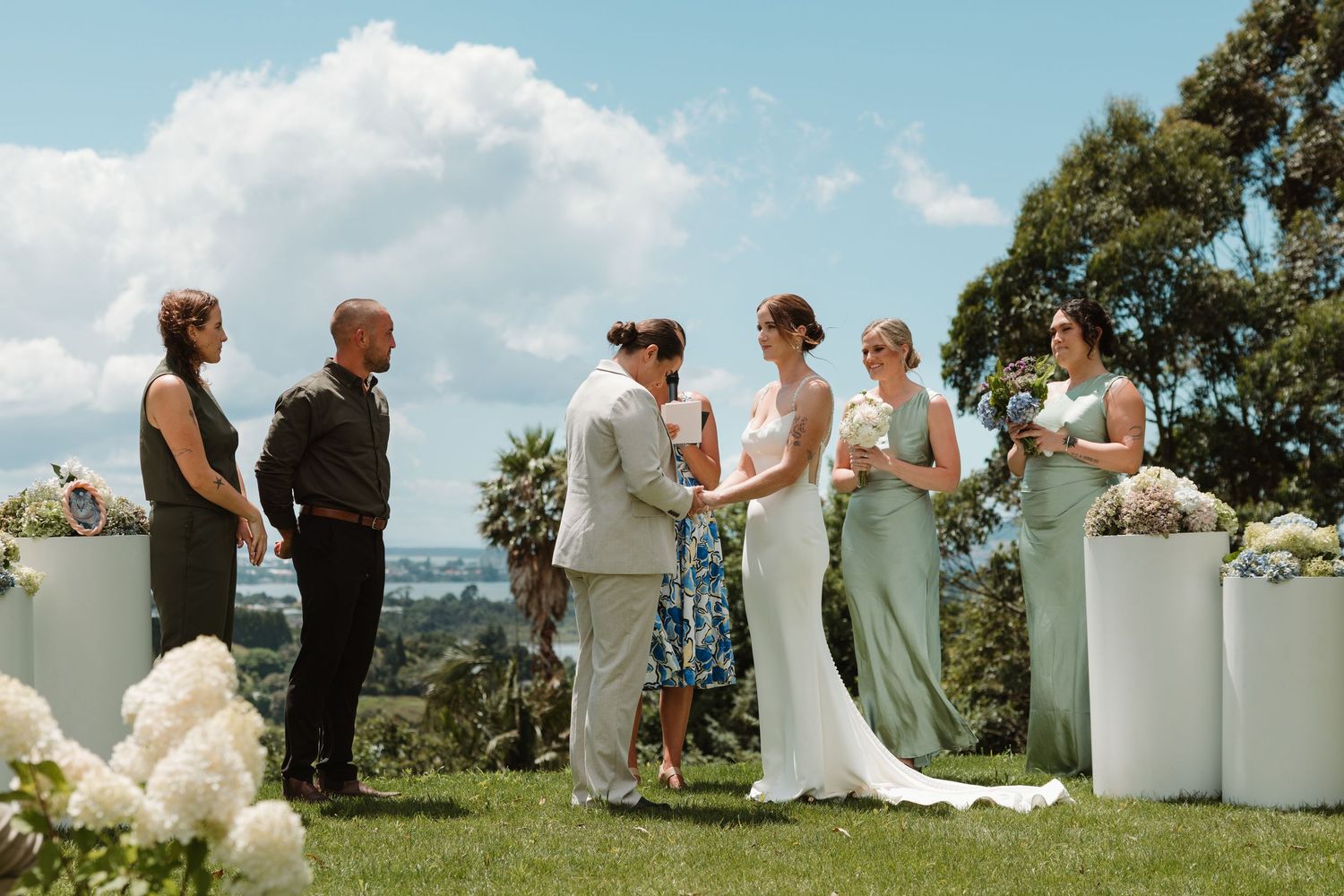 Outdoor wedding ceremony on green lawn with bride, groom, bridesmaids in sage green dresses, and guests under blue sky.