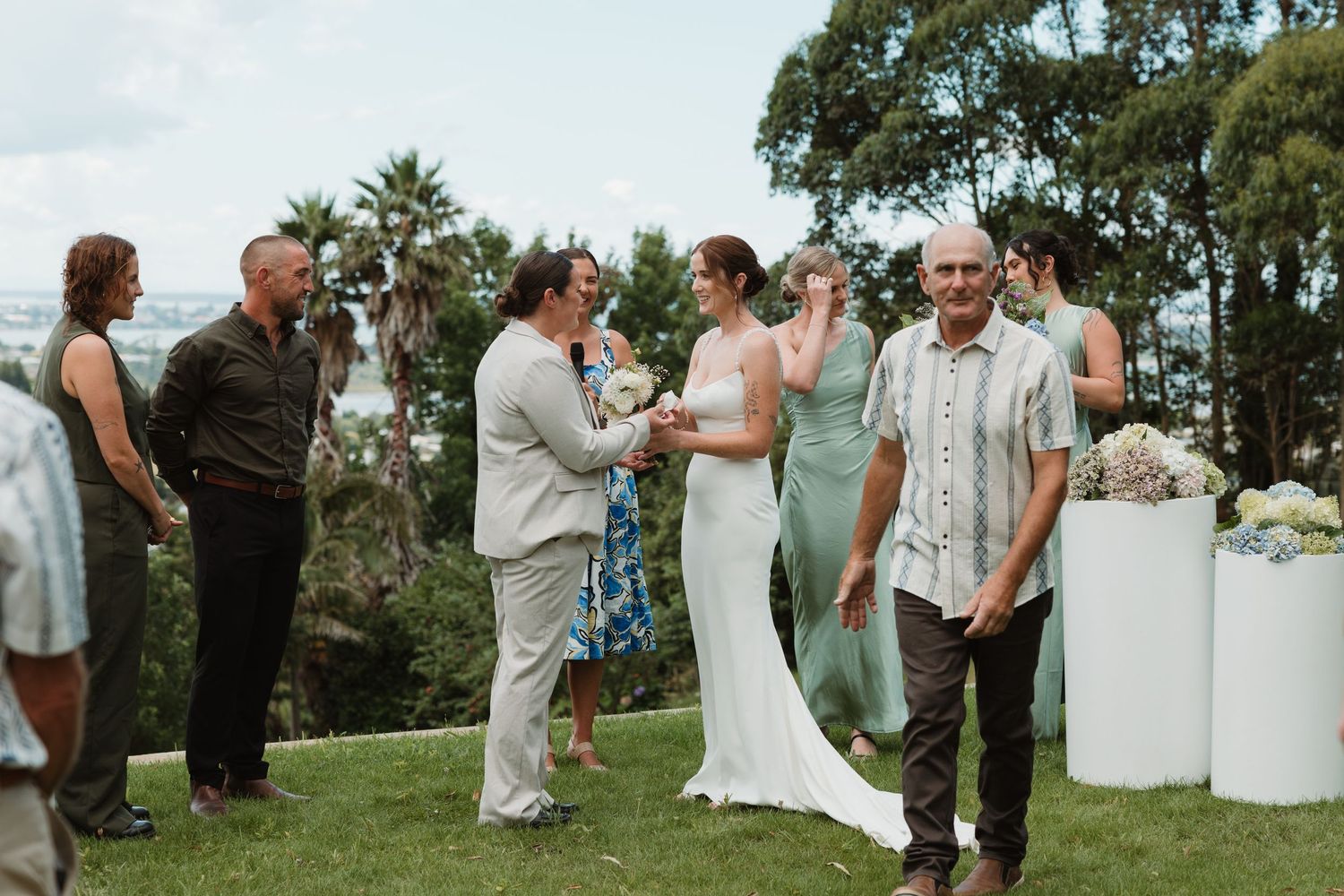 Outdoor wedding ceremony on green lawn with bride in white dress, groom, and guests surrounded by tropical trees.