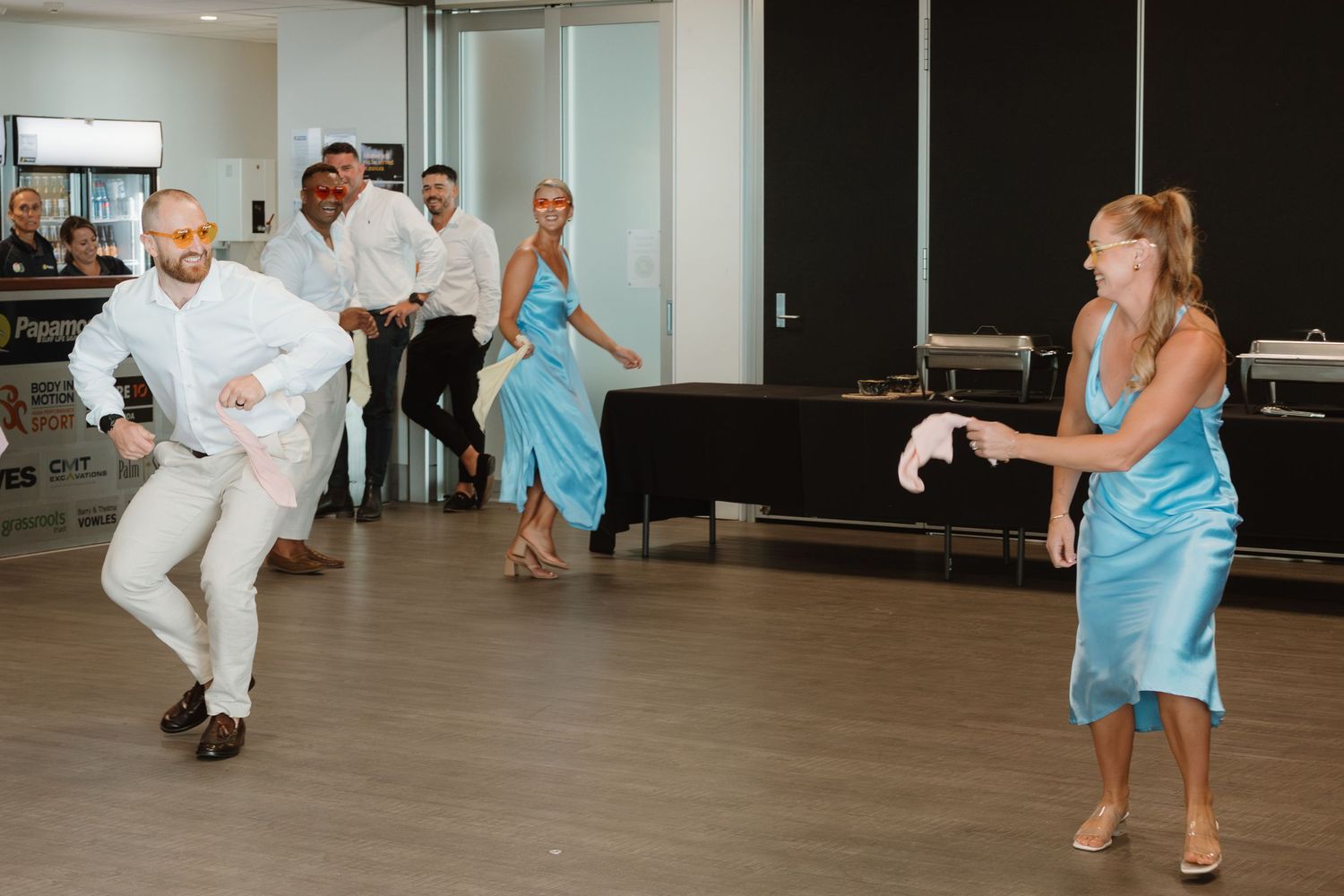 Wedding guests in formal attire dancing energetically at a reception venue, women in blue satin dresses.