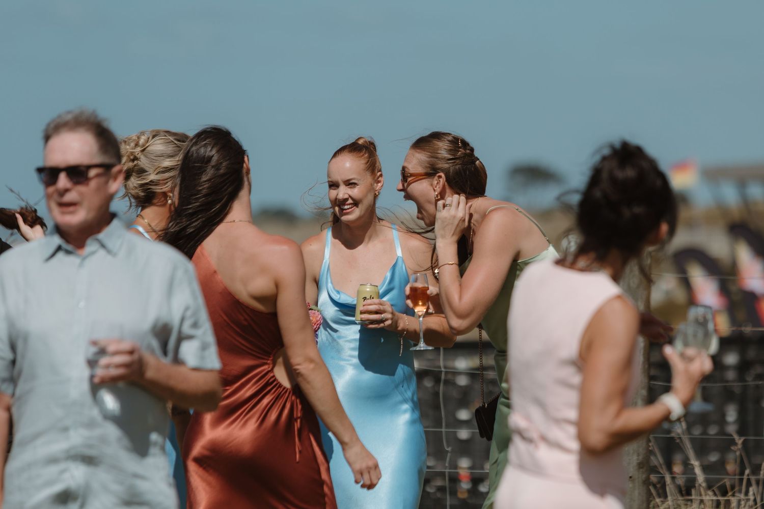Elegantly dressed women socializing outdoors at a summer event, holding drinks and laughing together.