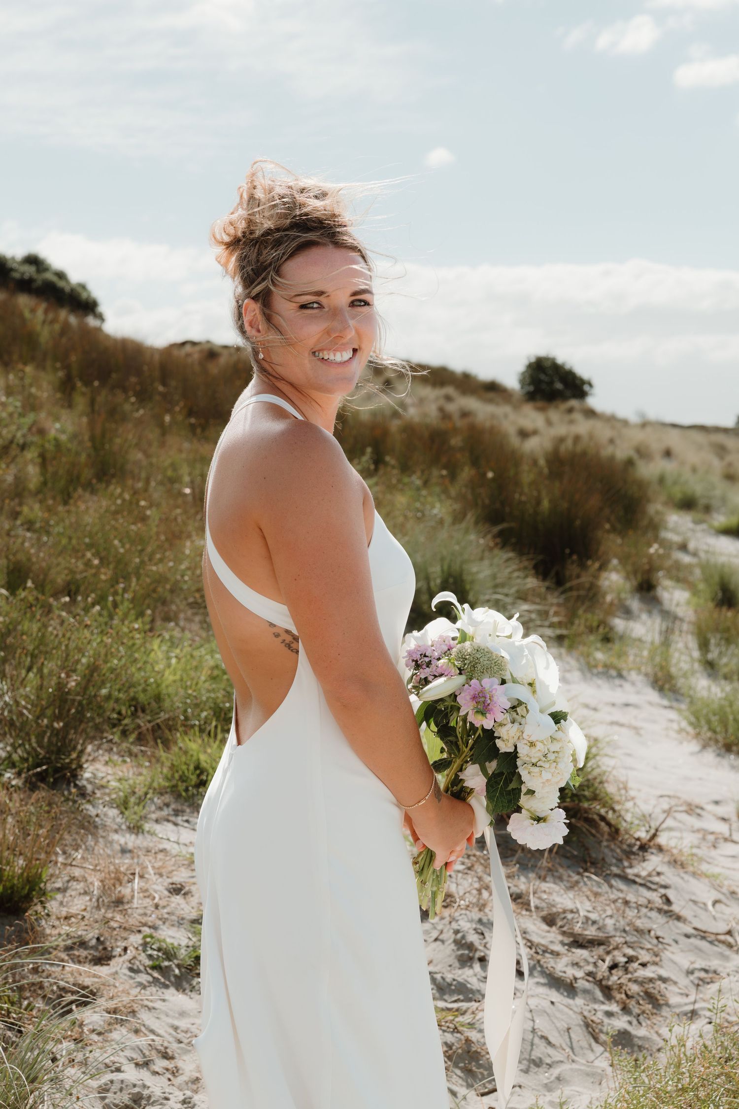 Smiling bride in white backless gown holding floral bouquet on sandy beach dunes on her wedding day.