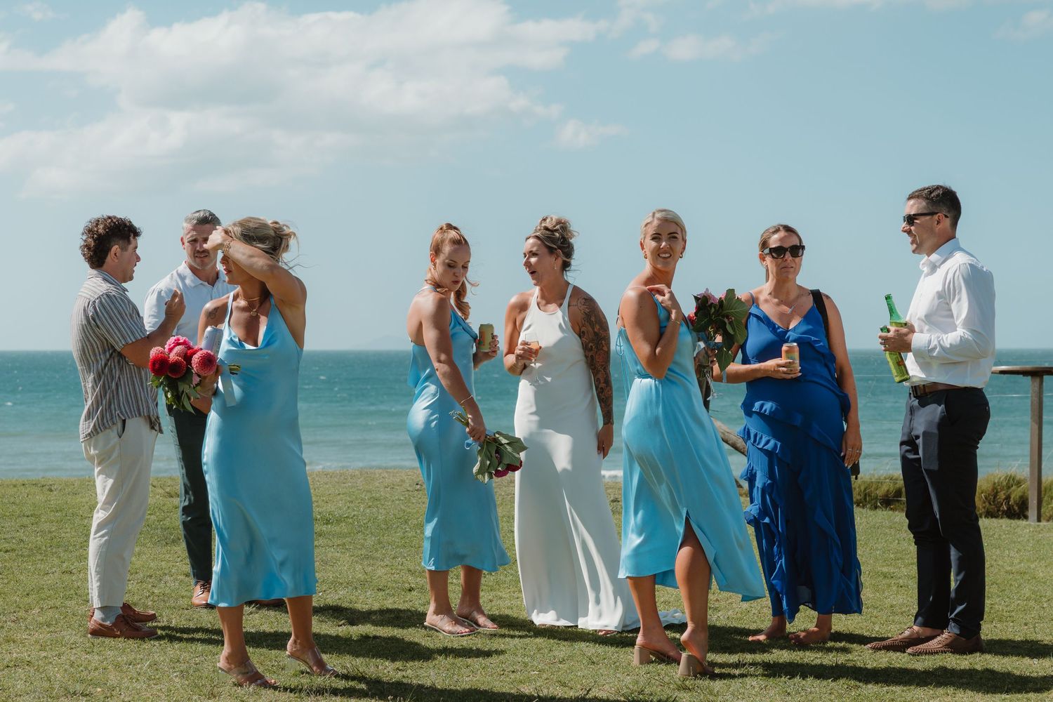 Bride in white gown with bridesmaids in blue dresses celebrating outdoors near ocean on a sunny wedding day.