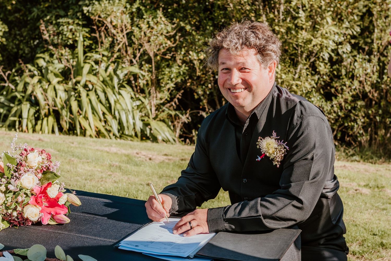 Someone signing documents outdoors while wearing formal attire and boutonniere.