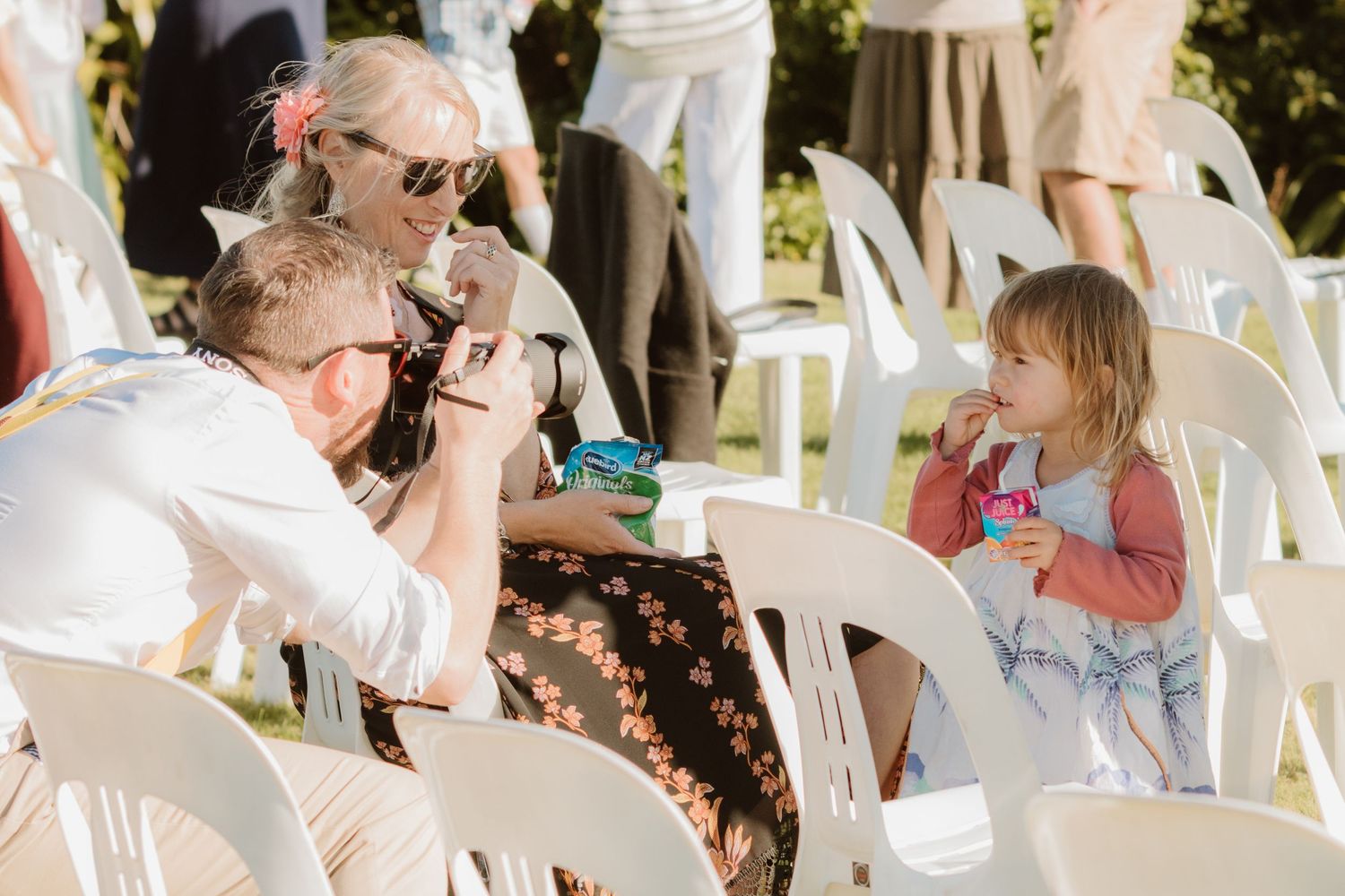 People enjoying drinks and conversation while seated in white chairs at an outdoor garden party event on a sunny day.