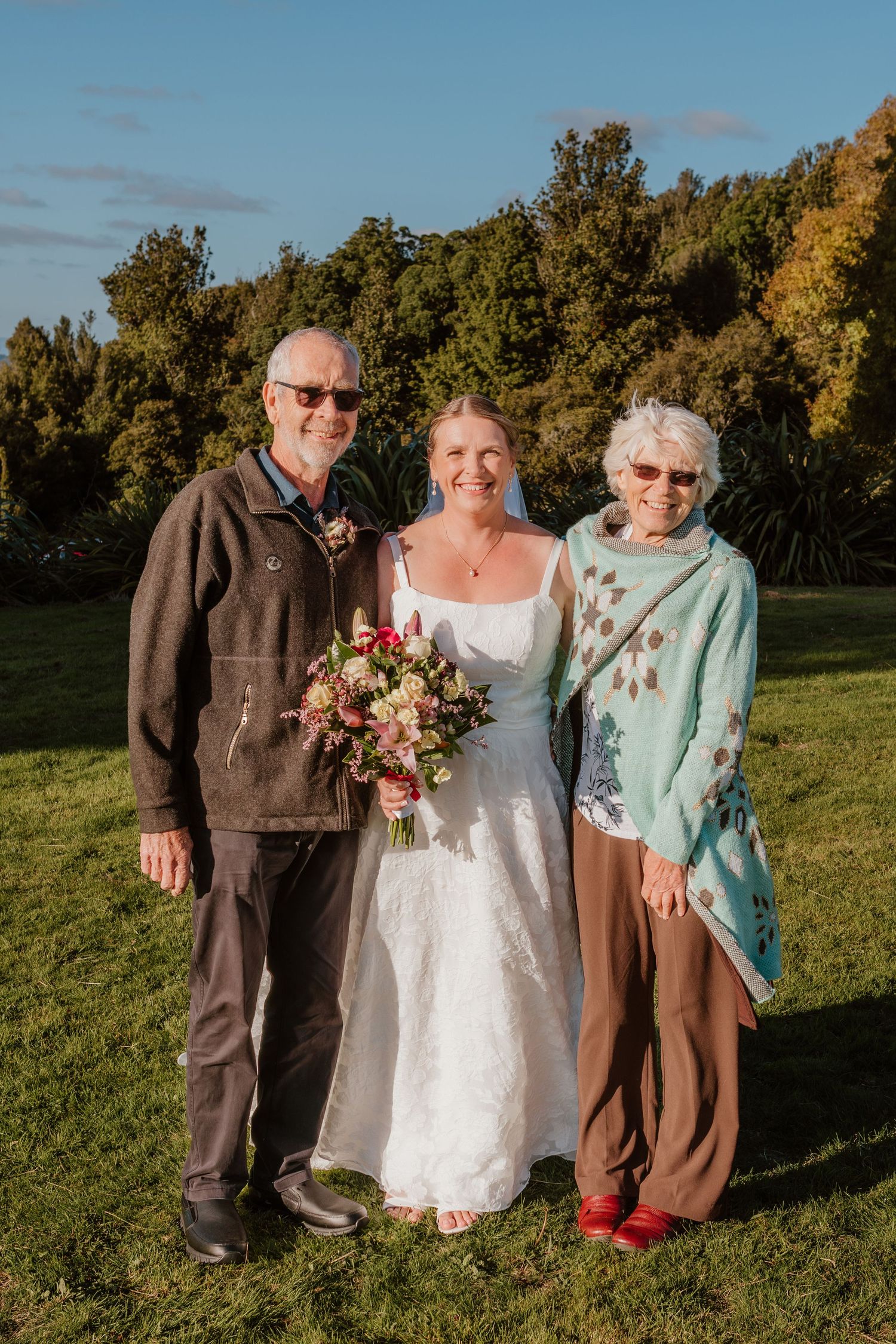 A wedding portrait outdoors on a sunny autumn day with three people posing together on grass with trees in background.