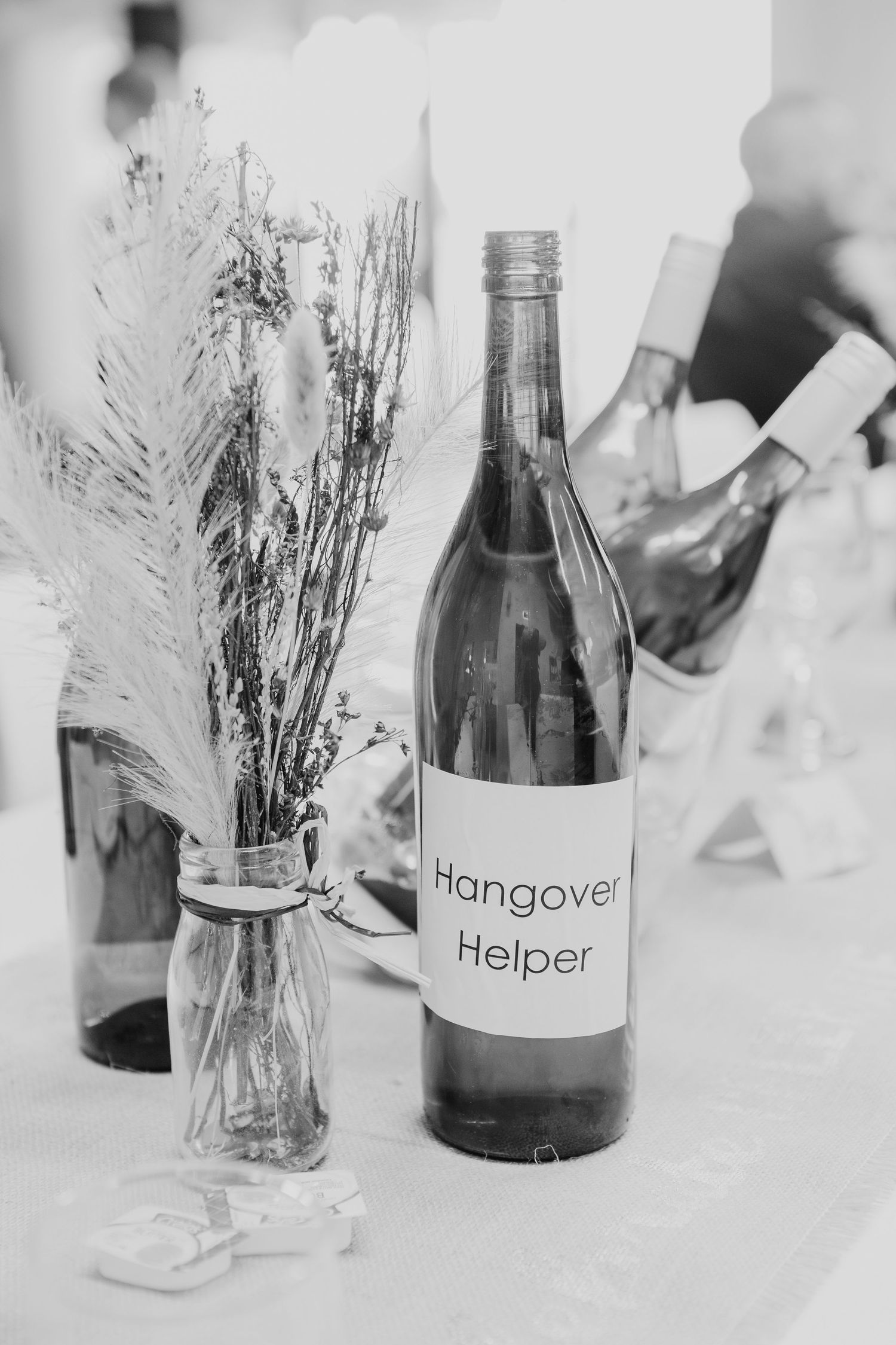 Black and white photo of a bottle labeled 'Hangover Helper' next to a vase with dried flowers on a table.