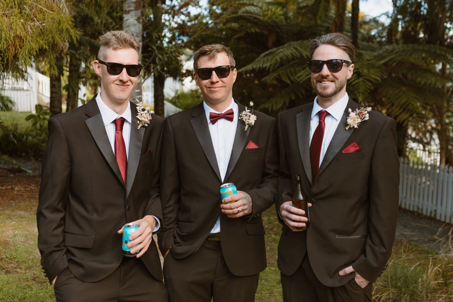 Three groomsmen in dark suits and sunglasses pose casually while holding drinks at outdoor wedding venue.