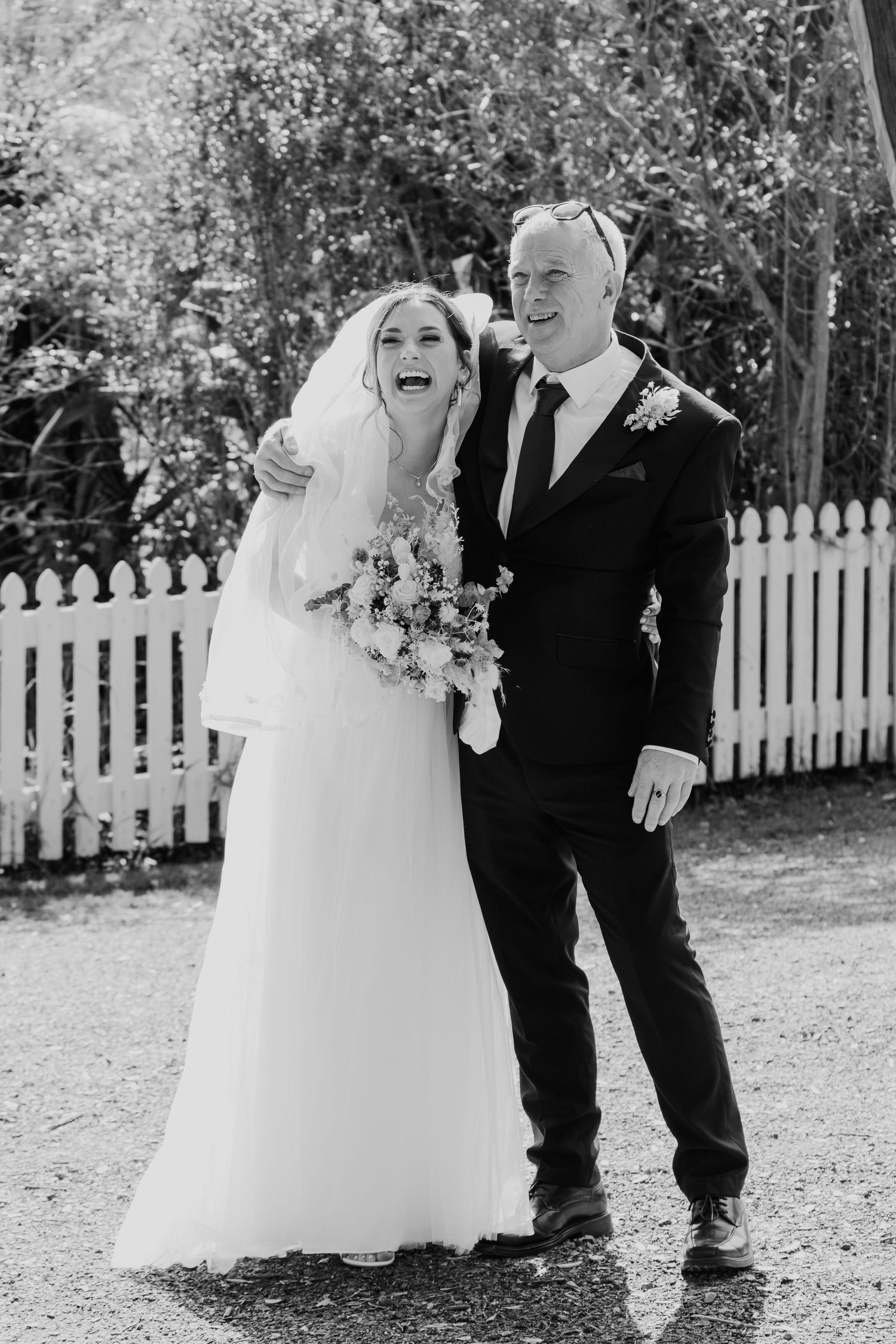 Black and white wedding photos show playful moments between a couple in formal attire near a white picket fence.