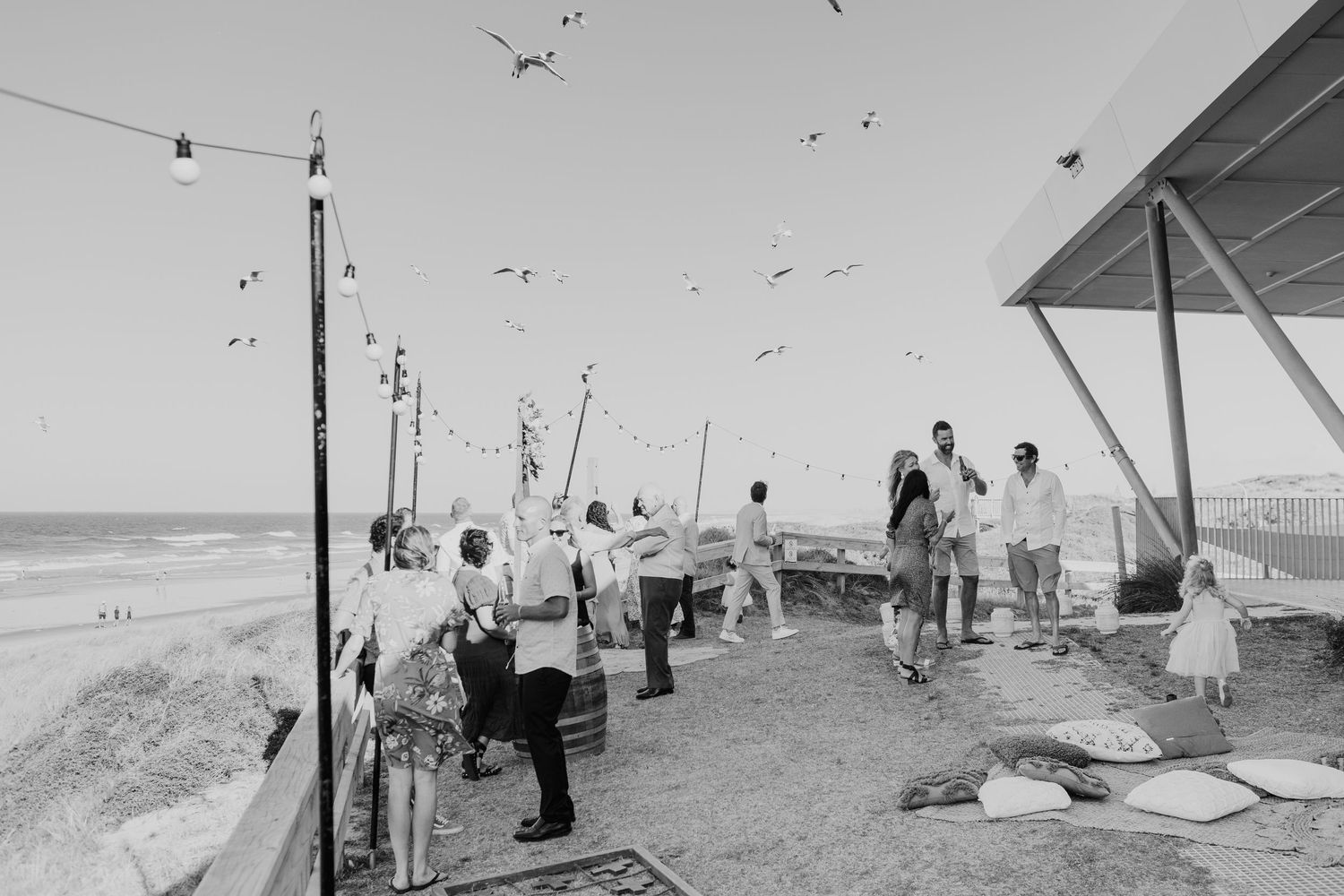 A black and white photograph shows people on a beach platform with seagulls flying overhead on a sunny day.