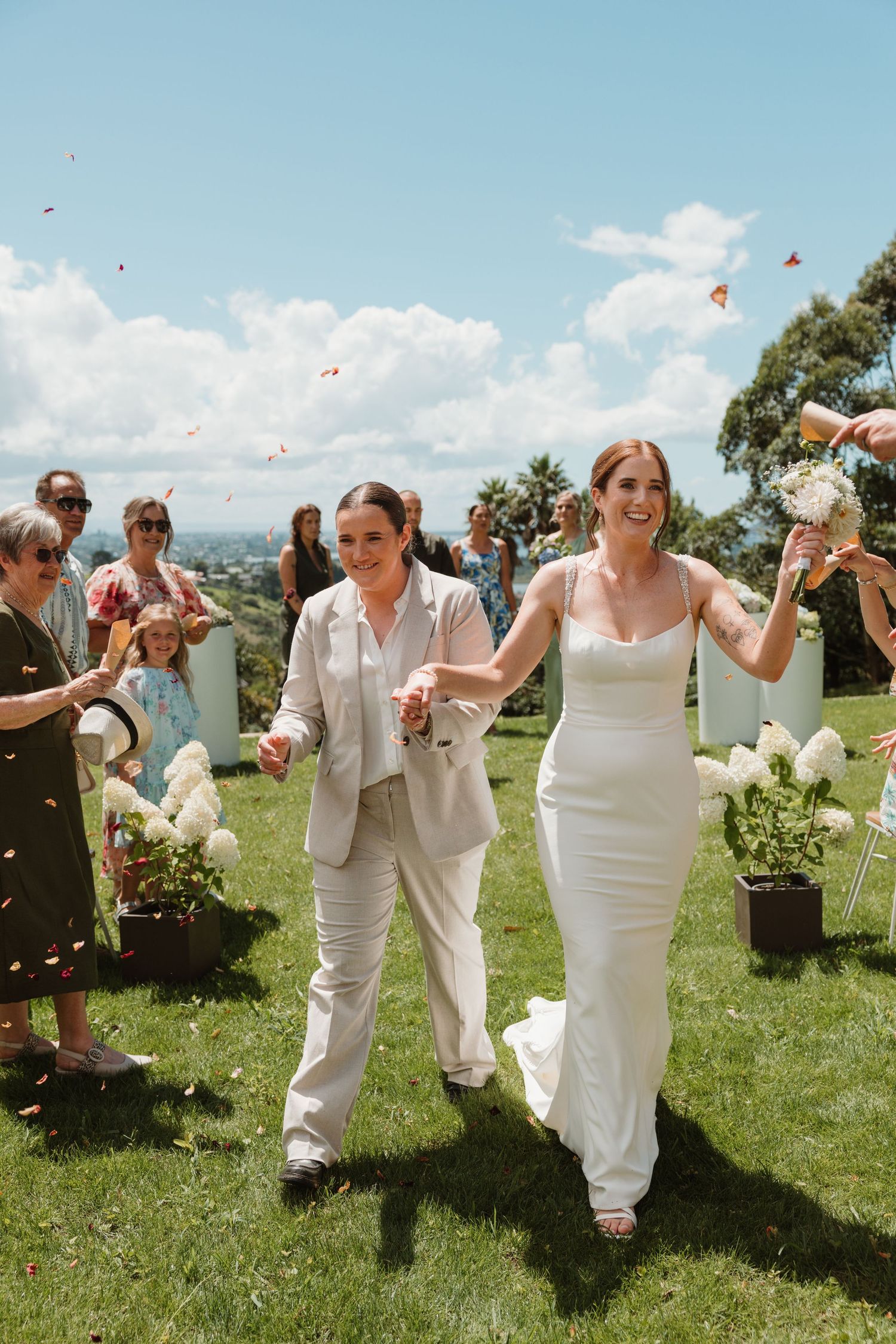 Wedding party celebrates on a sunny hillside by tossing flower petals into the blue summer sky.