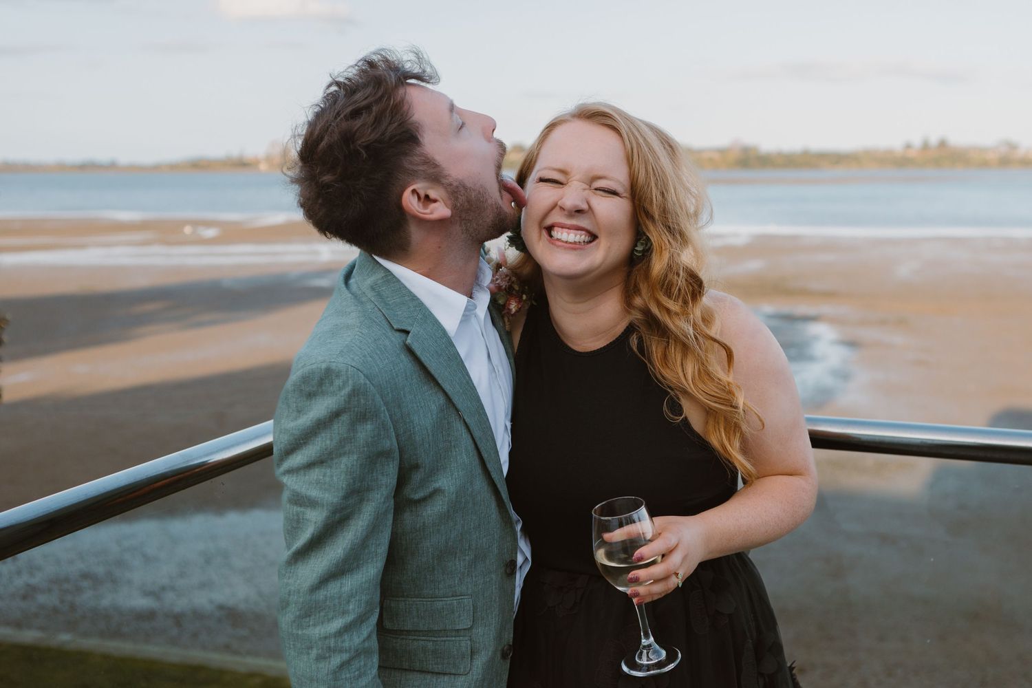 Romantic couple sharing wine and laughter during sunset beach photoshoot.