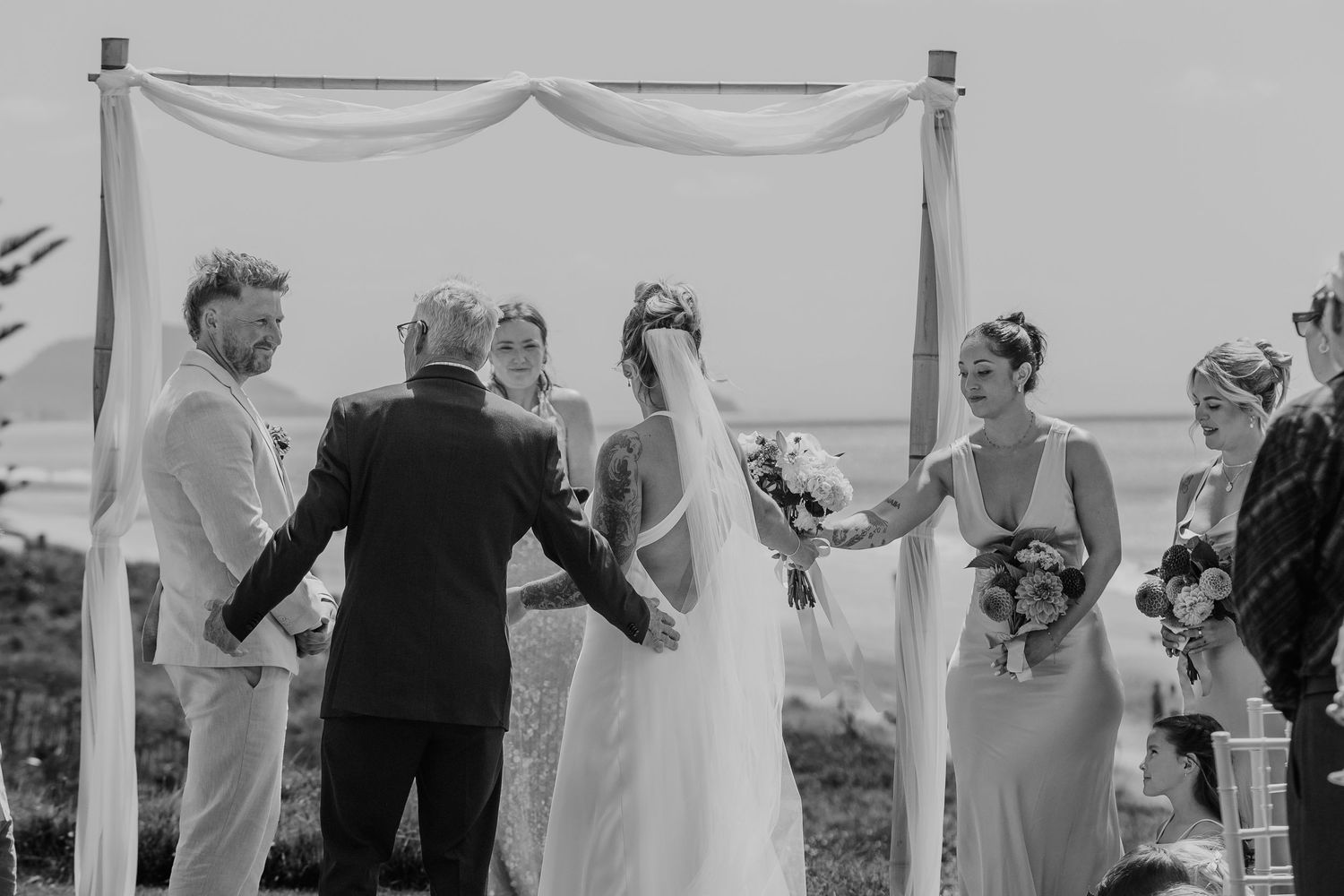Black and white outdoor wedding ceremony with bride, groom, officiant, and bridesmaids under a wooden arch by the ocean.