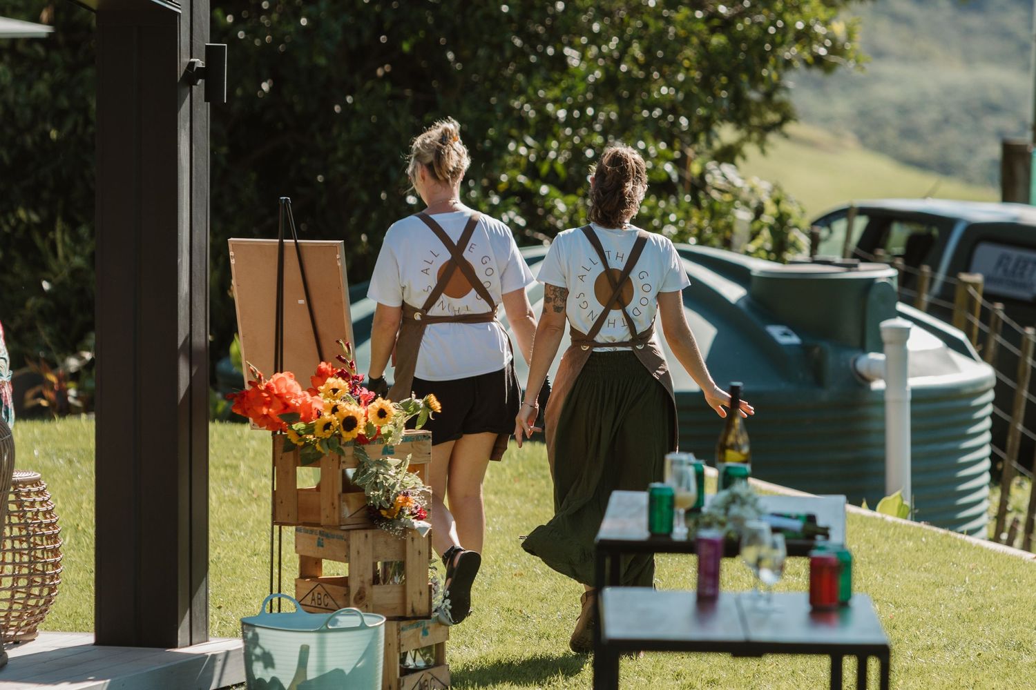 Two women in matching white shirts and suspenders walk through an outdoor market with flowers and rolling green hills.