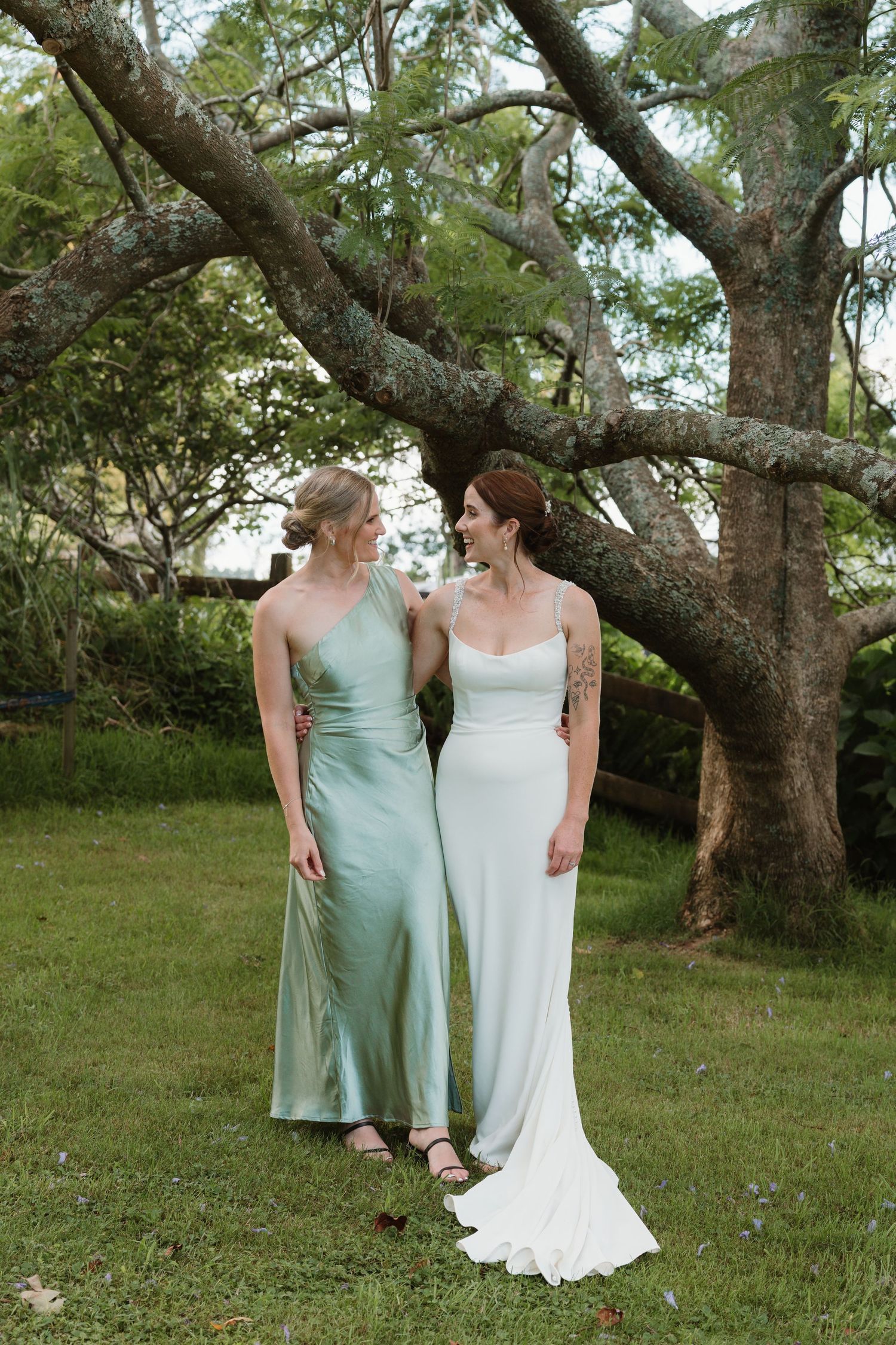 Bride in white gown and bridesmaid in sage green satin dress standing together on green lawn beneath large tree.