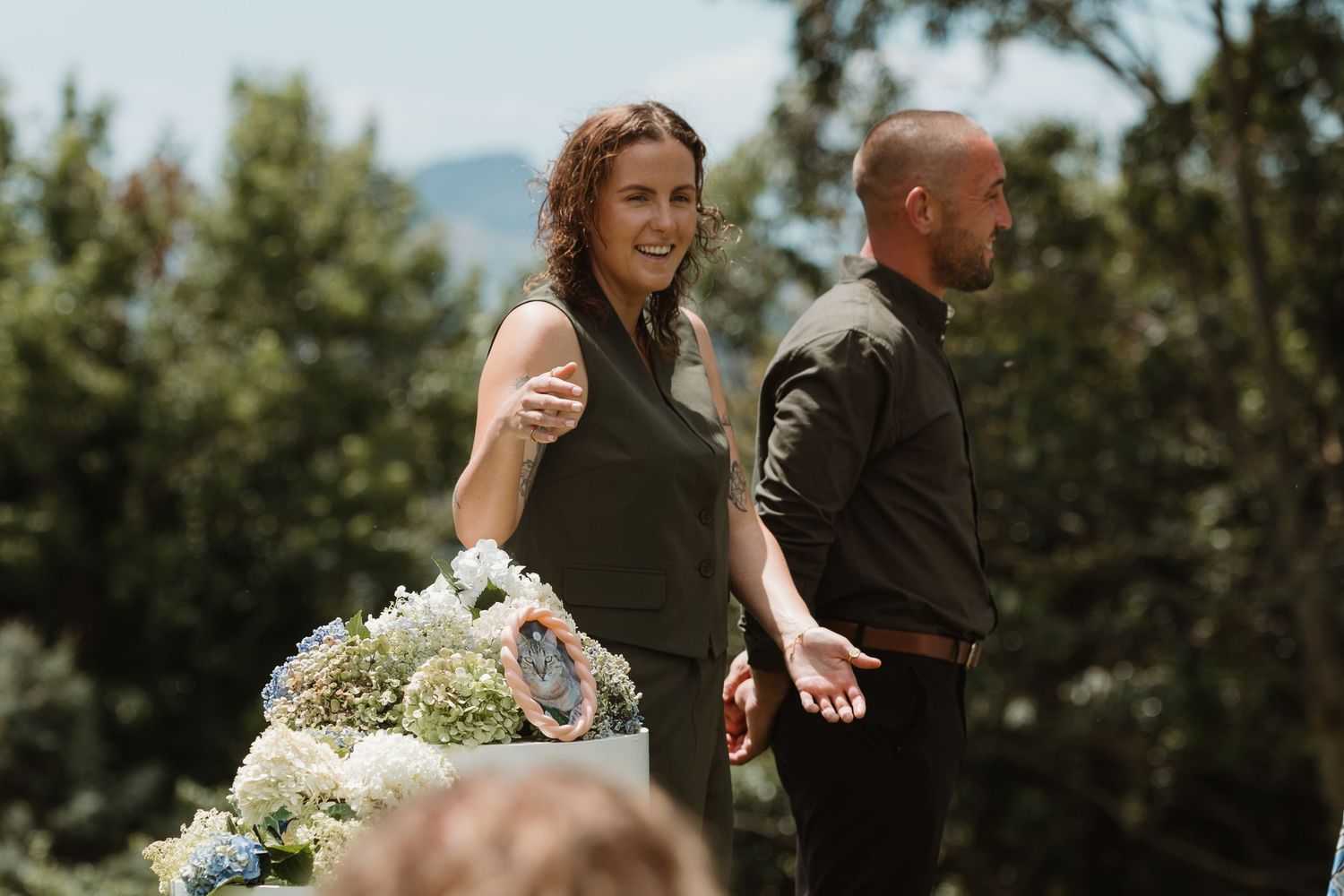 Woman in olive dress smiling outdoors at a floral arrangement with a man in black beside her.