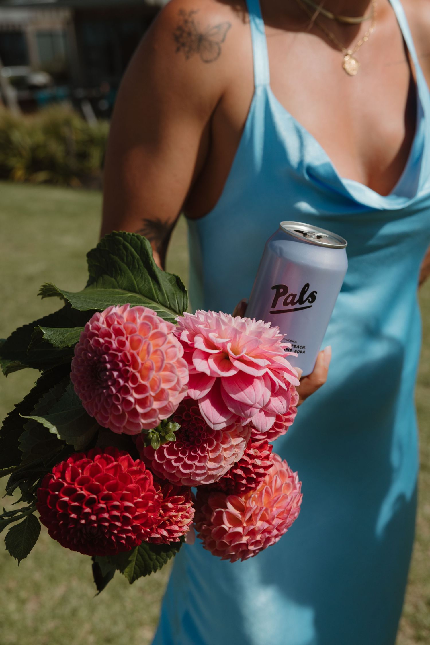 Woman in blue dress holding pink dahlia bouquet and Pals canned drink outdoors.