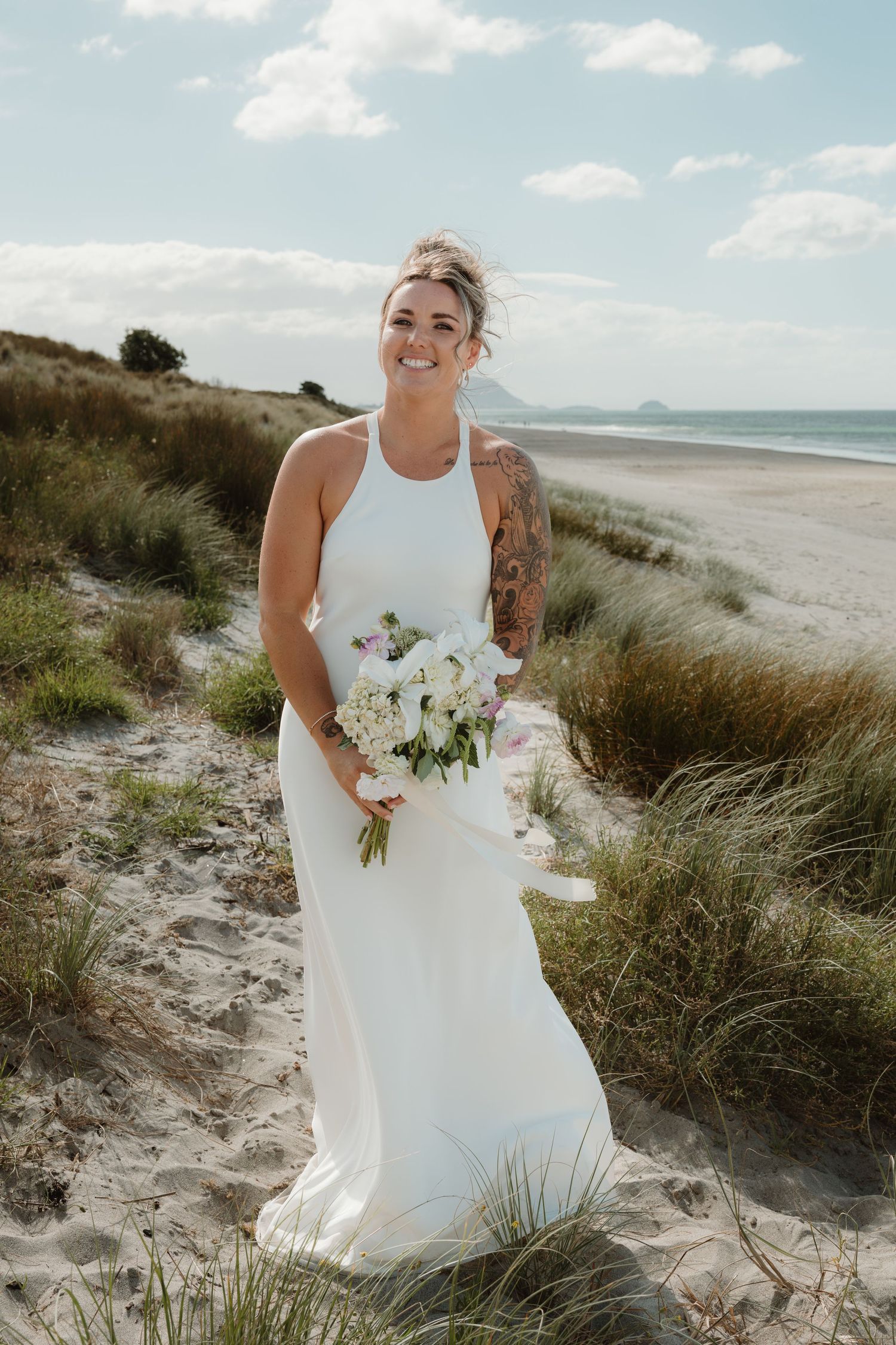 Smiling bride in white halter gown holding bouquet on sandy beach dunes with ocean backdrop.