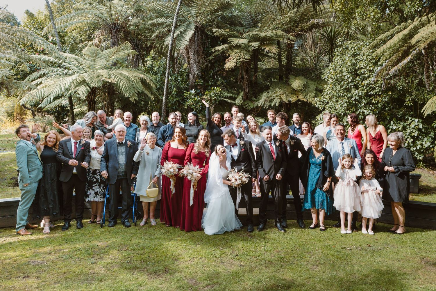 Large wedding party poses for a group photo in a lush garden setting with palm trees and greenery.