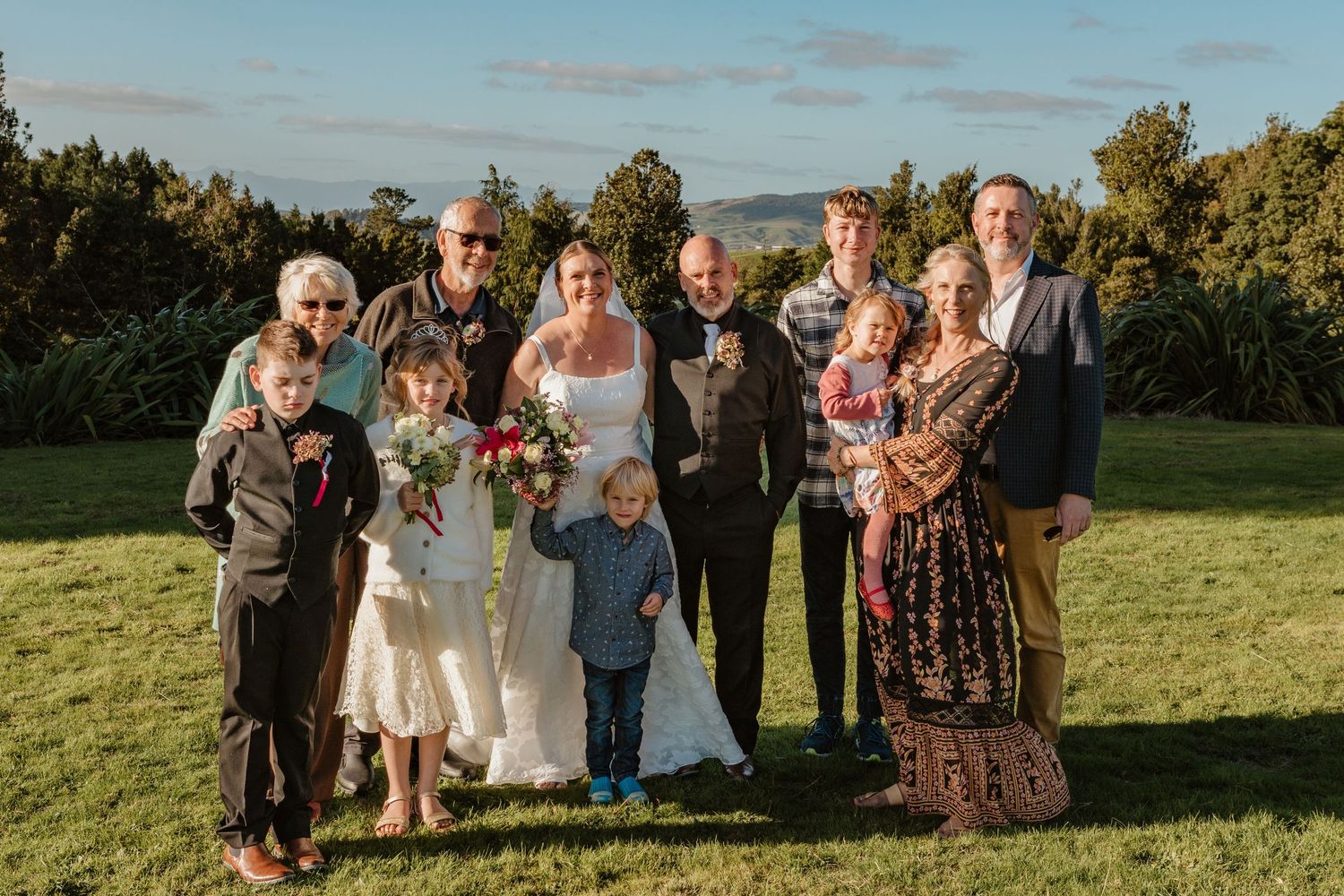 Wedding party poses together on grassy hilltop during golden hour celebration with trees and blue sky in background.