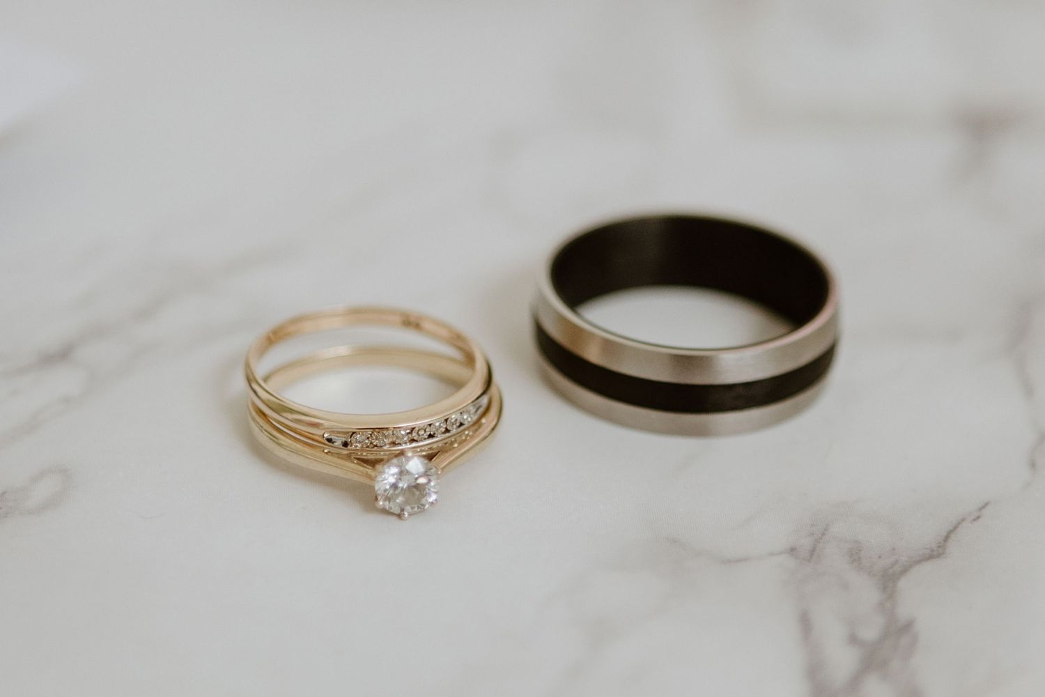 Two wedding rings on white marble surface - a gold engagement ring with diamond and a dark band with metallic detail.