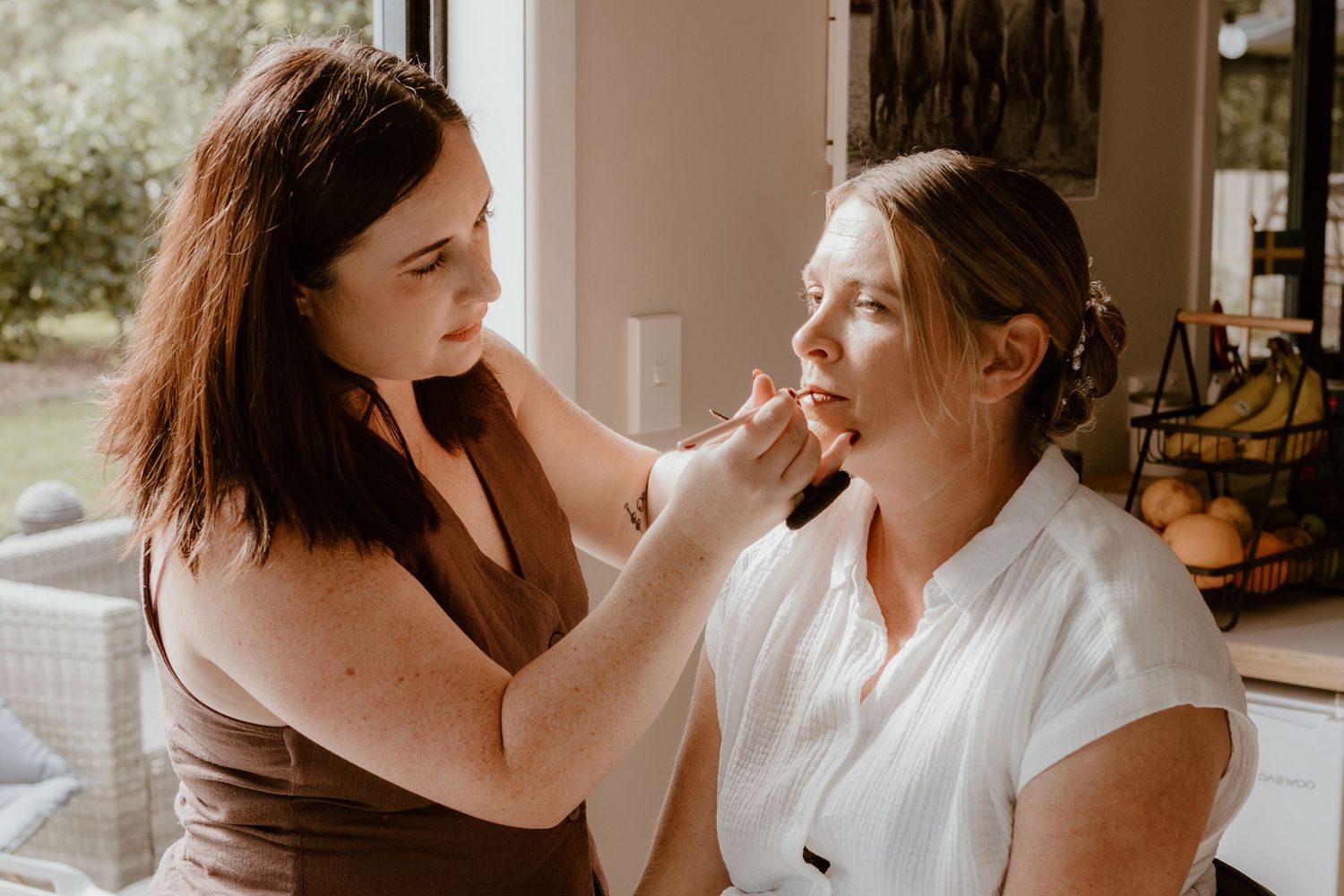 A makeup artist applies lipstick to a client in a naturally lit room during a beauty session.