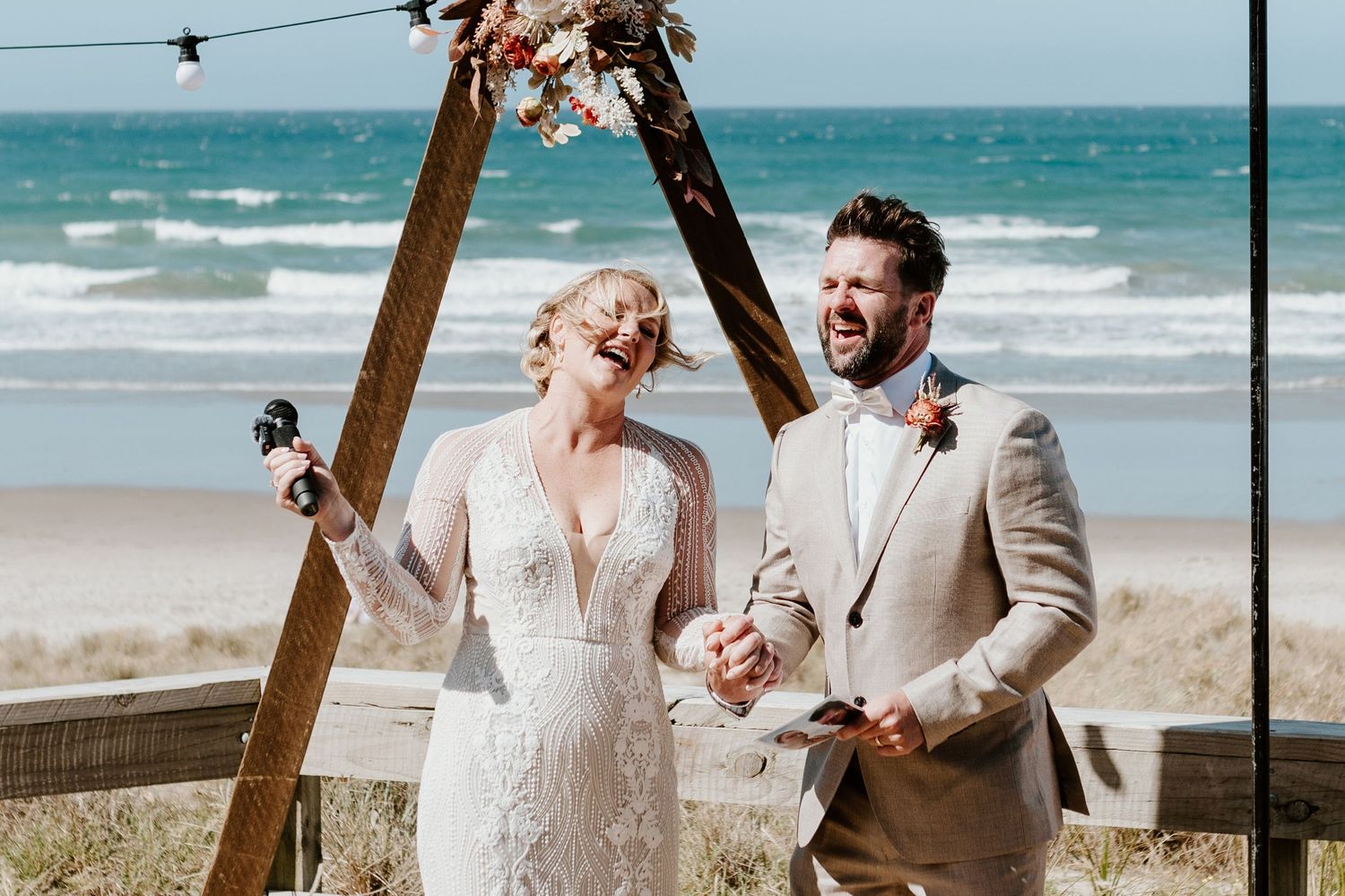 A joyful wedding couple celebrates under a wooden arch on a sunny beach with waves rolling in the background.