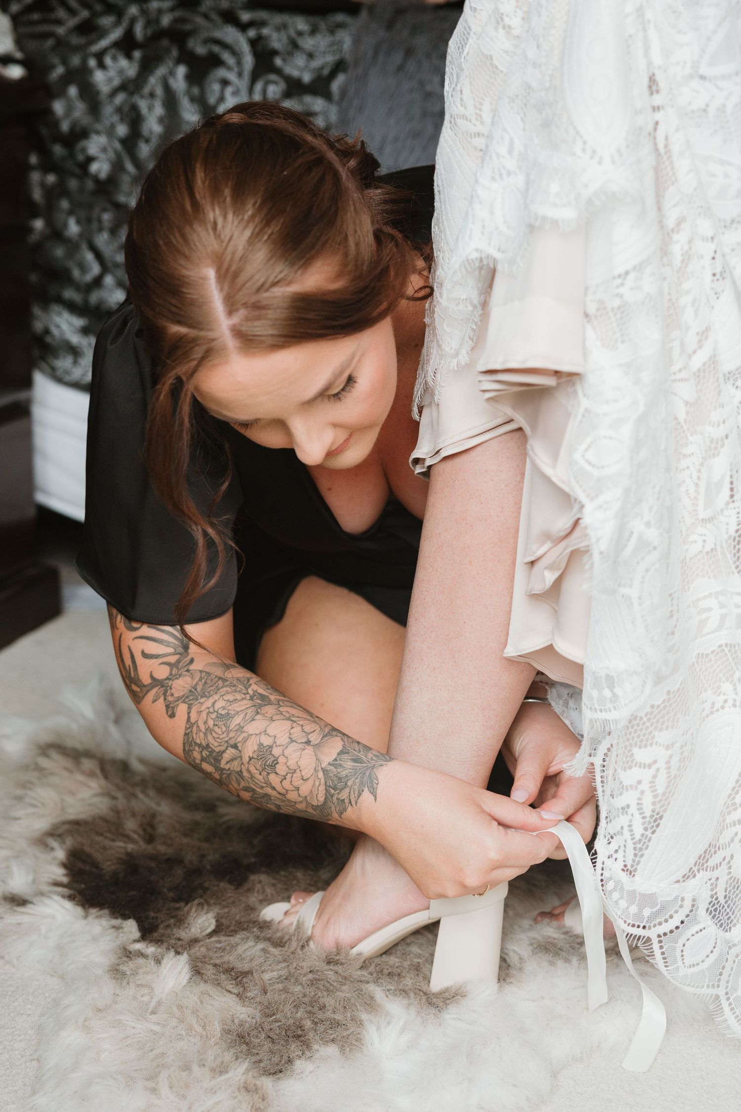 A person in a white lace dress adjusts their shoe while sitting on a grey furry rug.