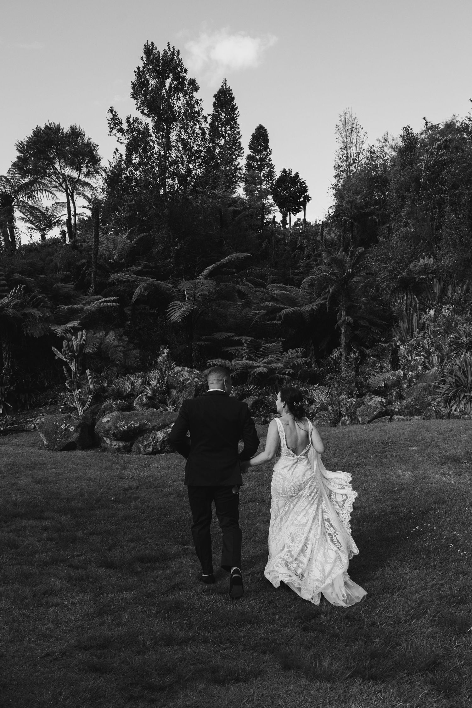 A couple in formal wedding attire walks away from the camera across a garden lawn in black and white.