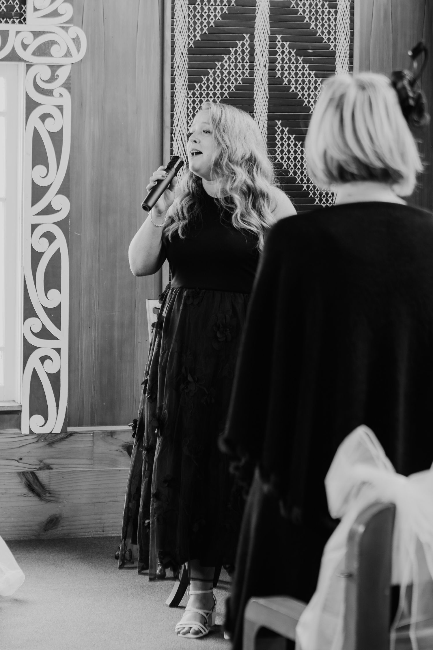 Singer performing at wedding ceremony in black dress against ornate white backdrop.